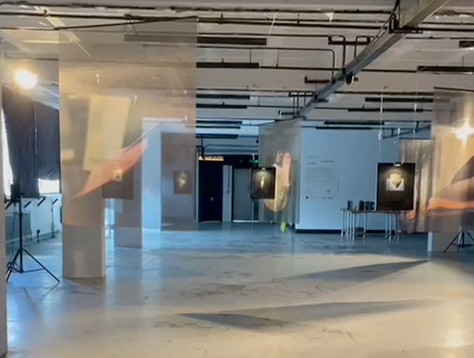 Empty gallery or exhibition space with artworks displayed on walls and hanging from ceiling, with lighting and some light reflections.