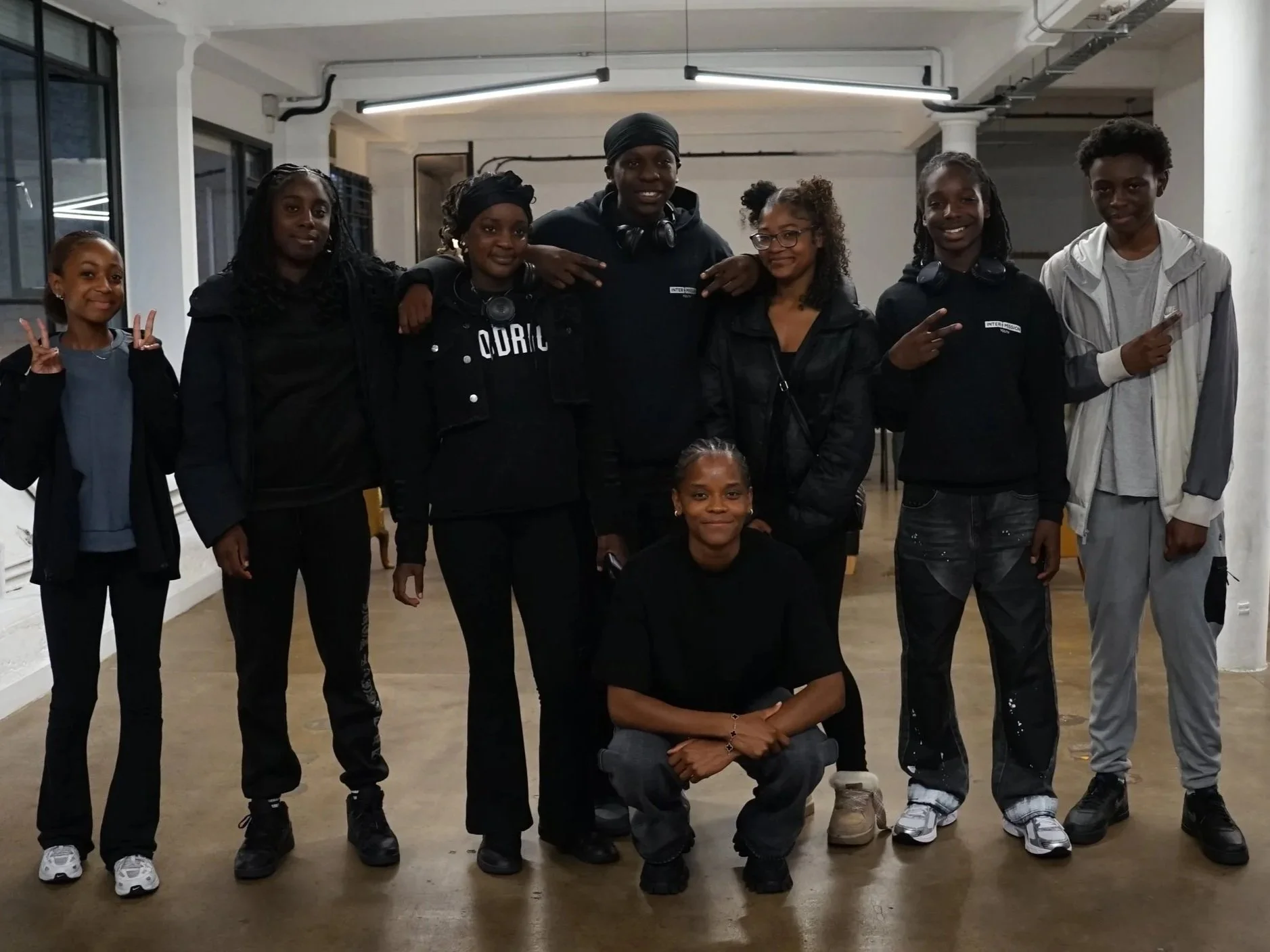A group of seven young people and one adult woman standing indoors in a well-lit room, smiling and posing for the camera. Some are making peace signs, and one person is sitting in front of the group. They appear to be in a casual setting.