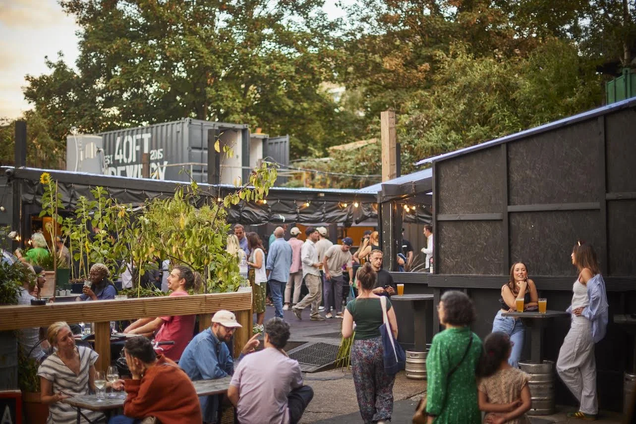 People socializing and enjoying drinks at an outdoor rooftop bar with string lights and lush green trees in the background.