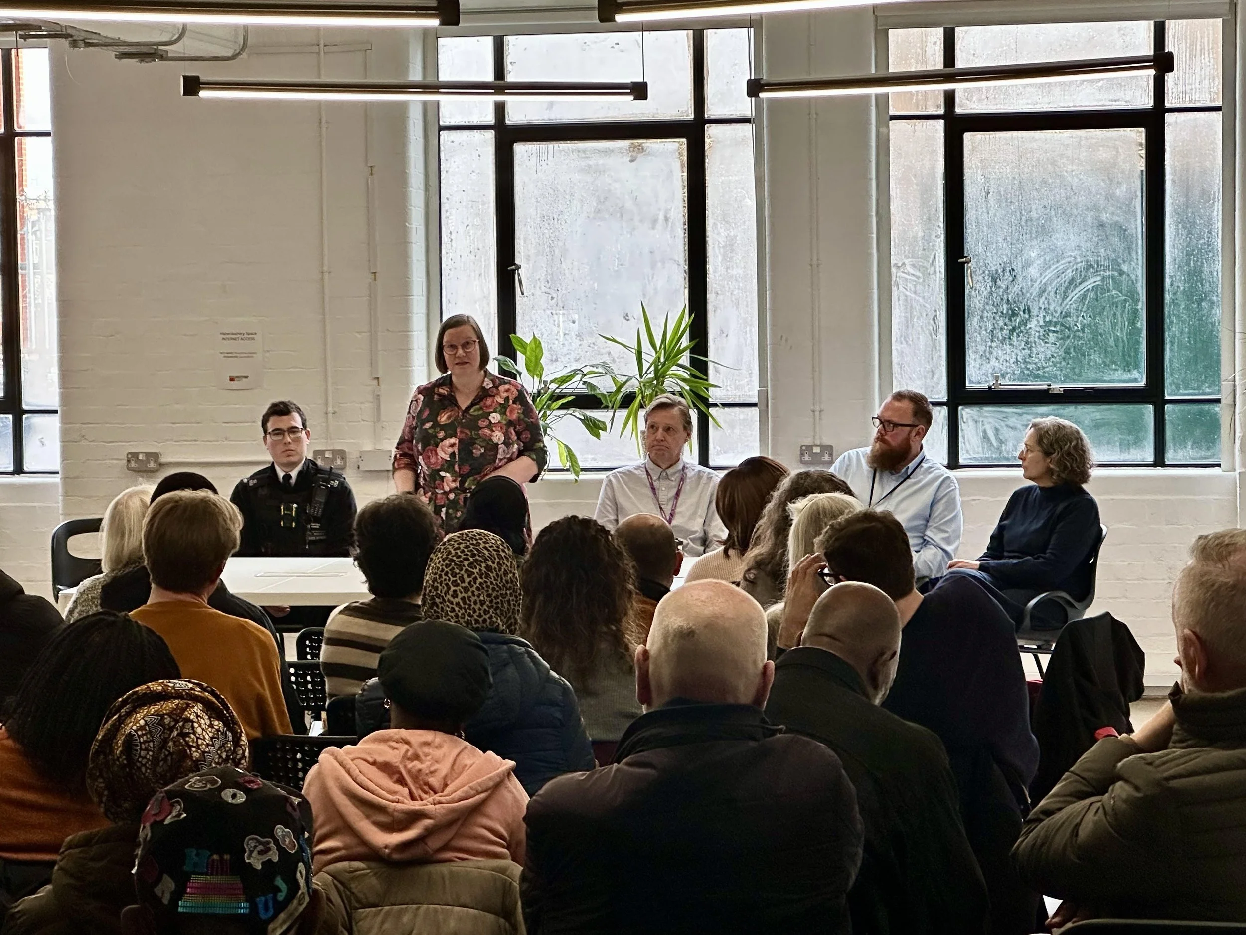 A woman wearing glasses and a floral shirt stands in front of a seated group of people, speaking during a presentation or discussion in a large room with big windows, white brick walls, and a potted plant.