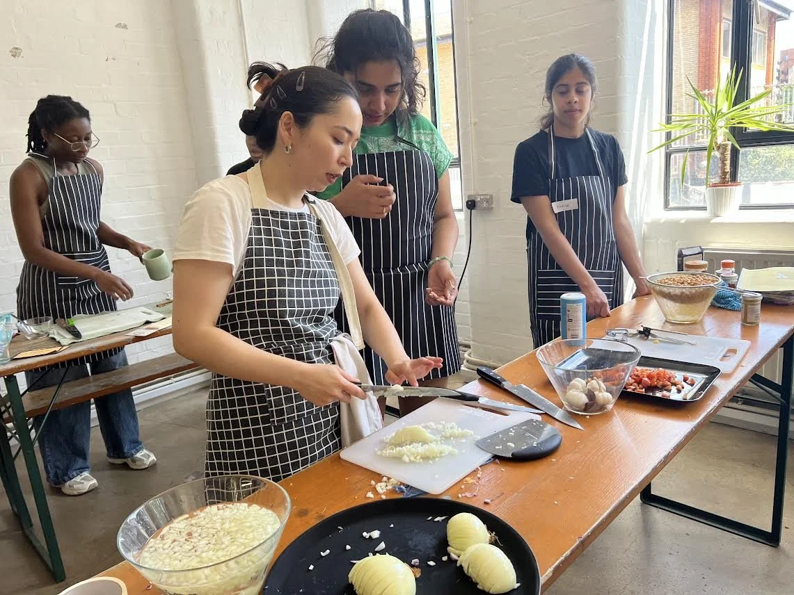 Group of five women cooking and preparing ingredients in a kitchen with natural light and a large window, some chopping vegetables, wearing aprons.
