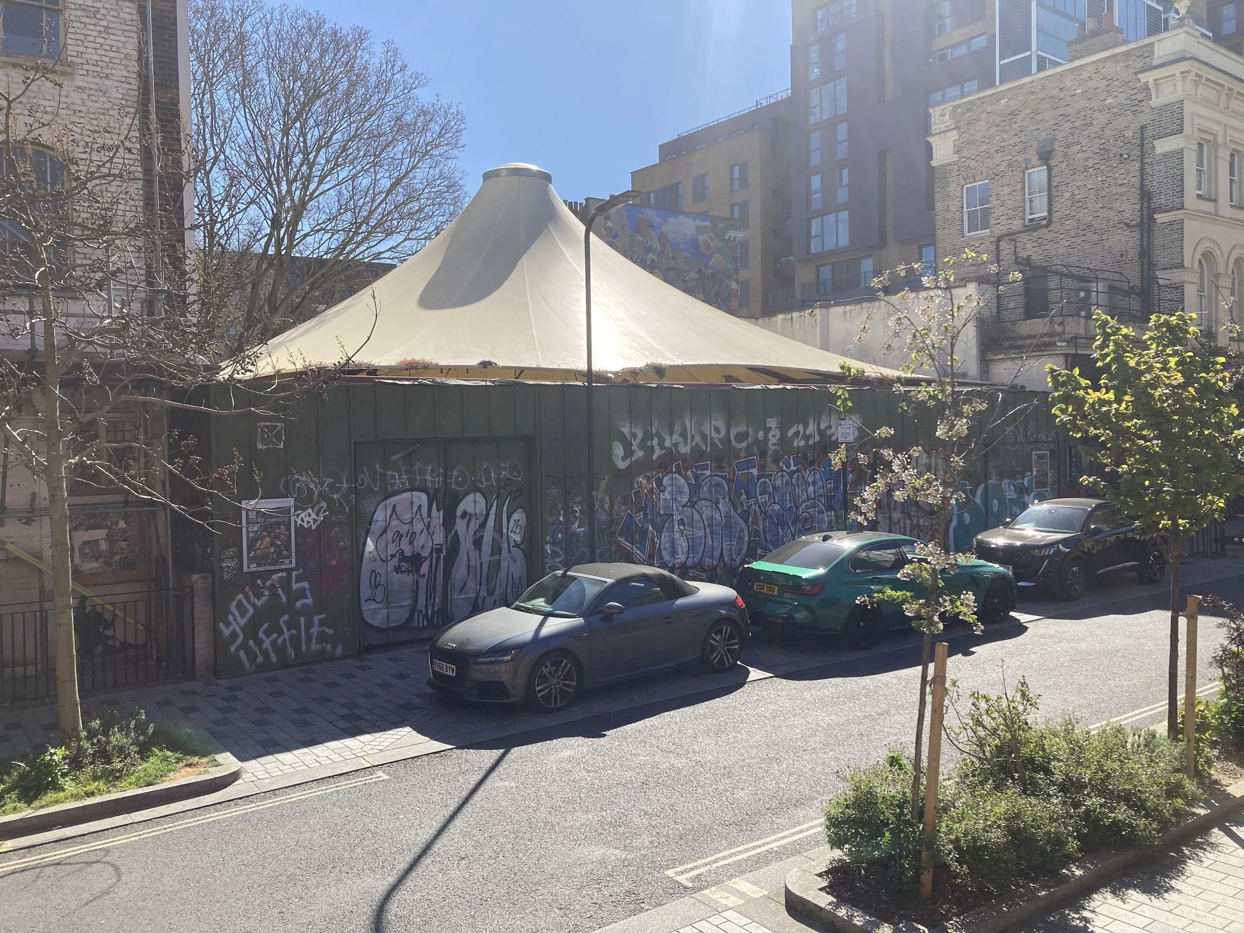 A street scene with parked cars, graffiti-covered wall, and buildings in the background. Trees without leaves are visible on the left and right sides.