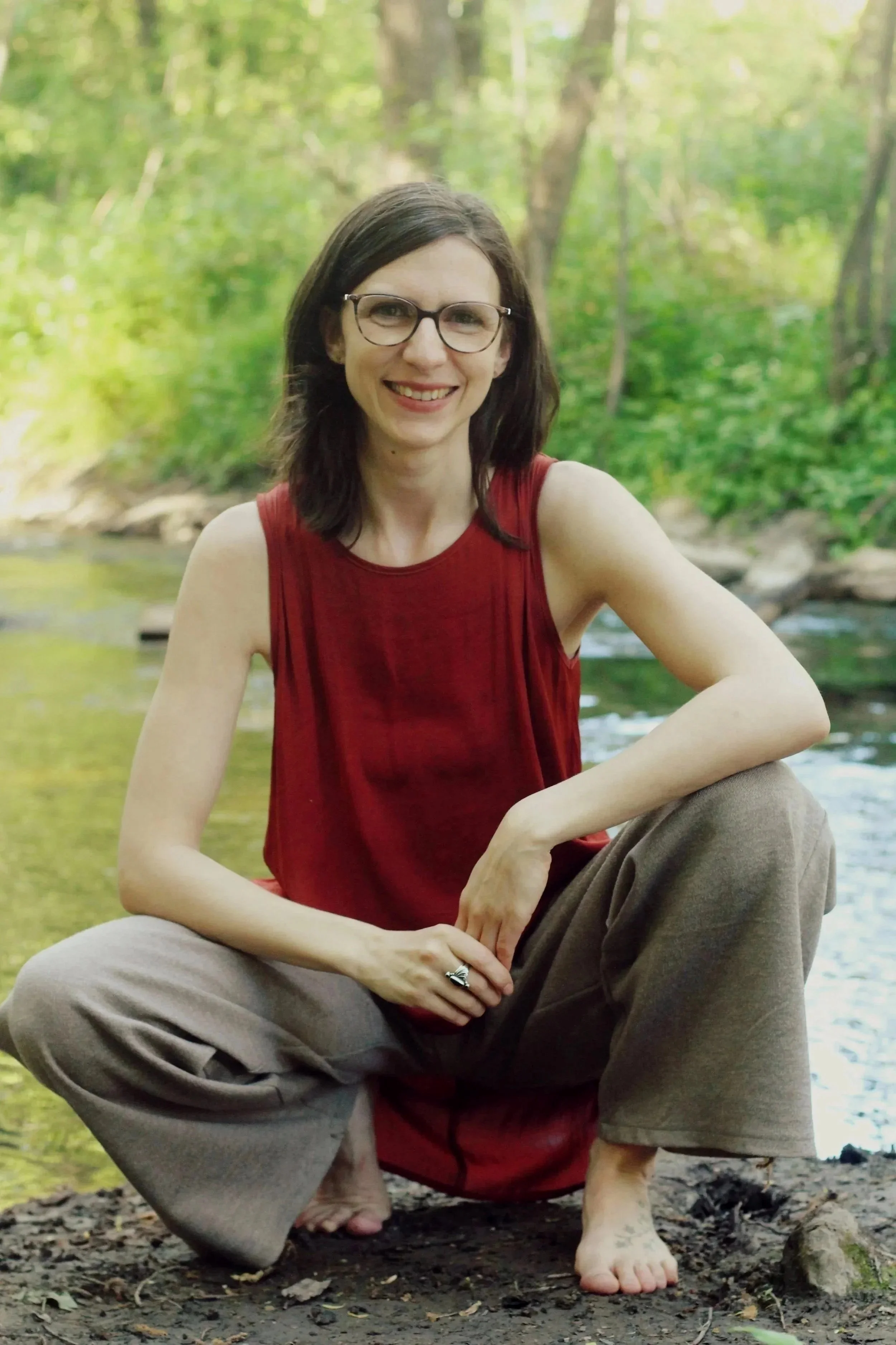 A woman with glasses and dark hair, wearing a red sleeveless top and brown pants, is crouching by a river in a forested area, smiling at the camera.