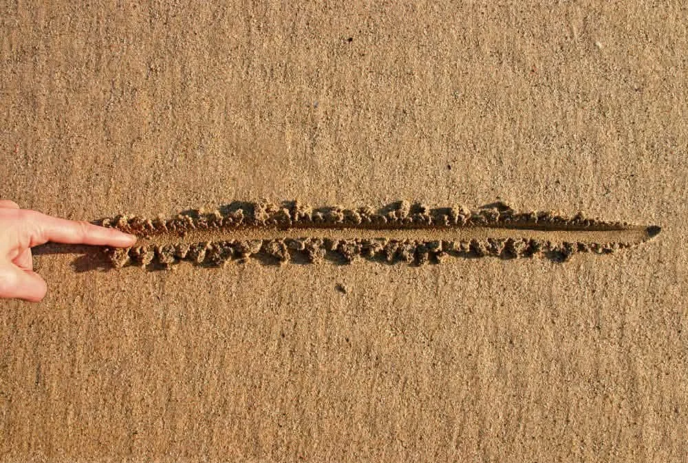 Close-up of a pointed stick dragging a line in the sand at the beach, with a hand, and a clear sky.
