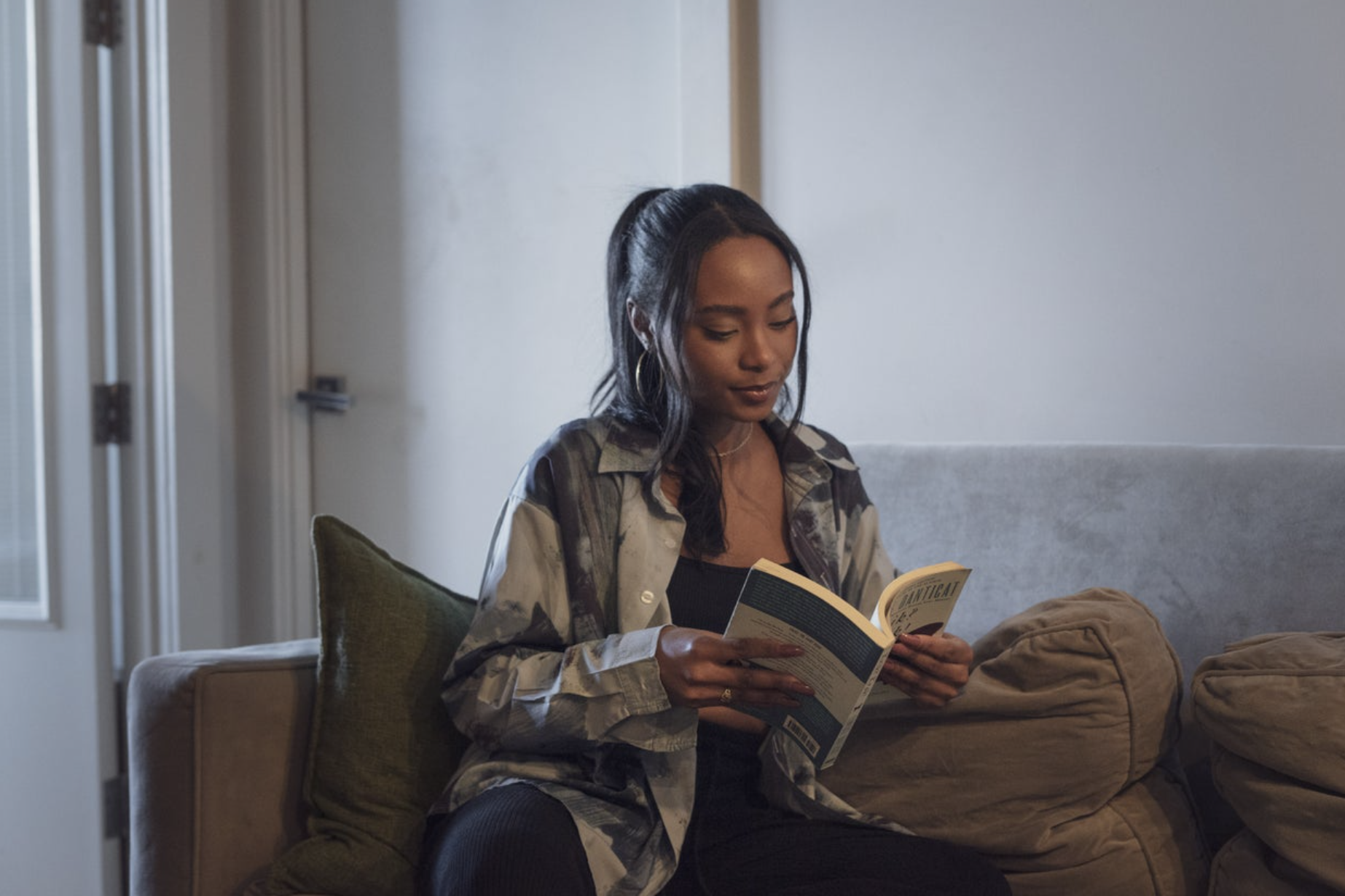 Young woman with long dark hair in a high ponytail, wearing hoop earrings and a camouflage jacket, sitting on a beige sofa with a green pillow, reading a book indoors.