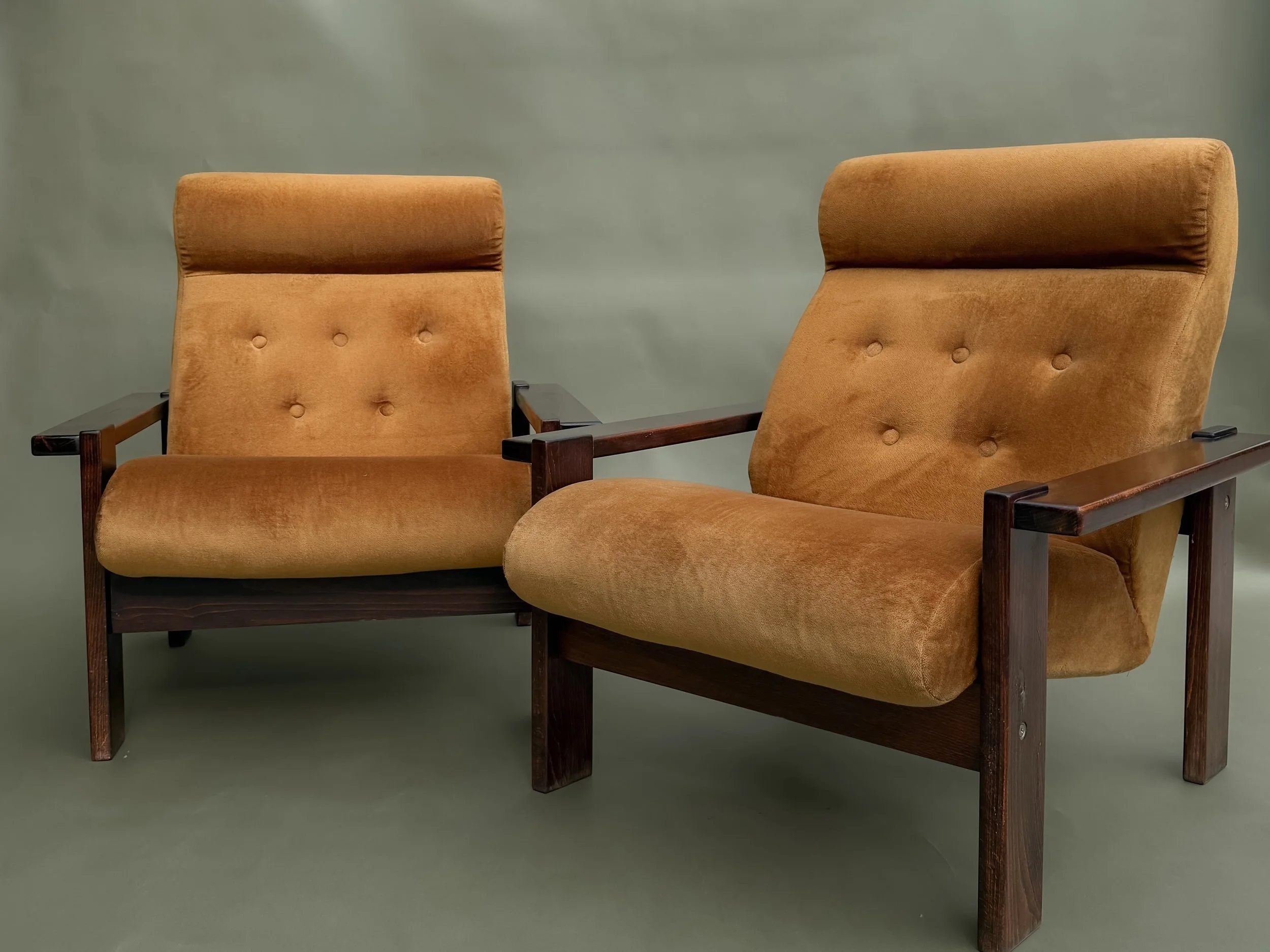 Two vintage-style armchairs with wooden arms and legs, upholstered in mustard-colored velvet fabric, set against a plain background.