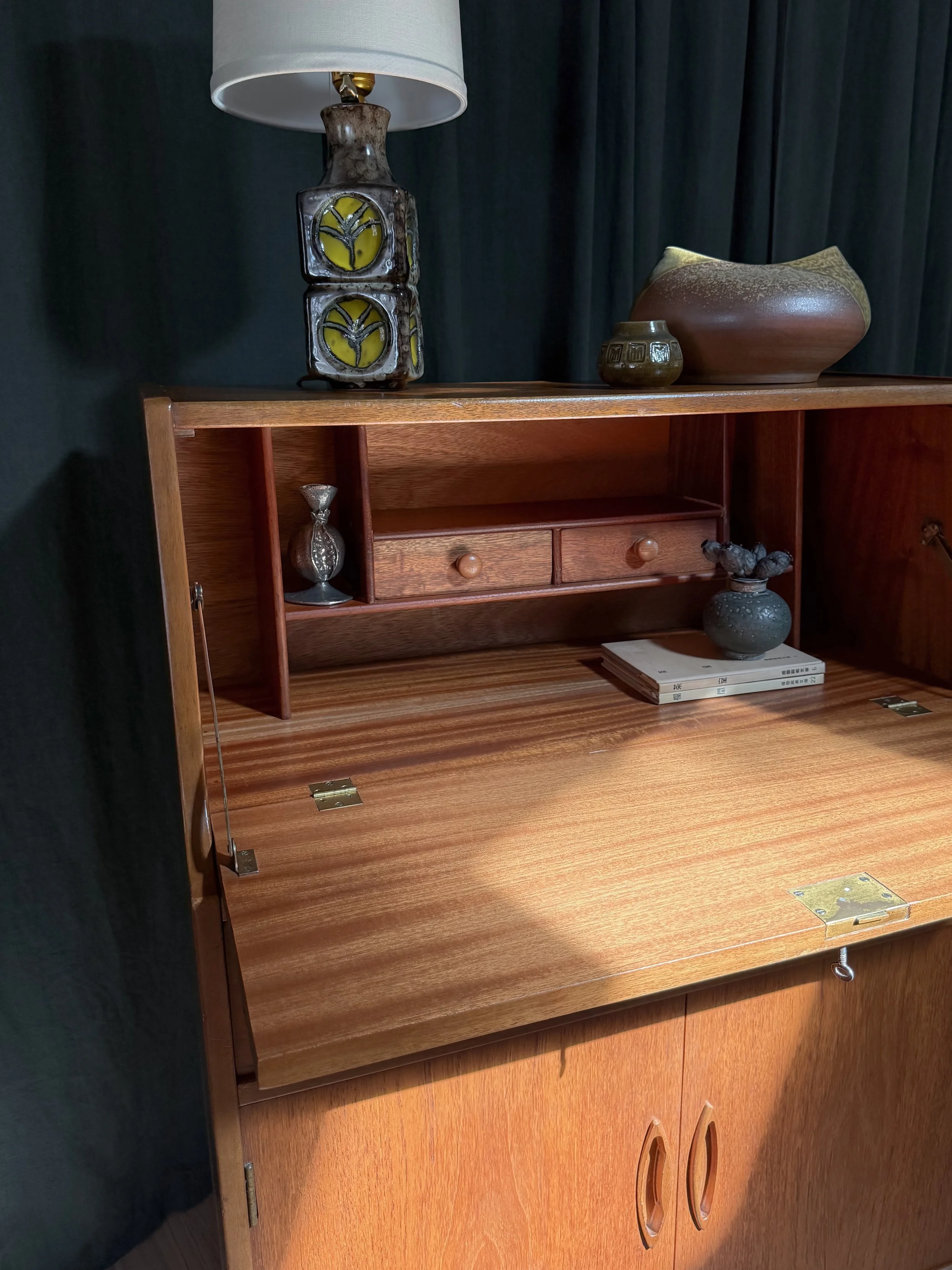 Wooden secretary desk with decorative vases and books on top, and a dark curtain background.
