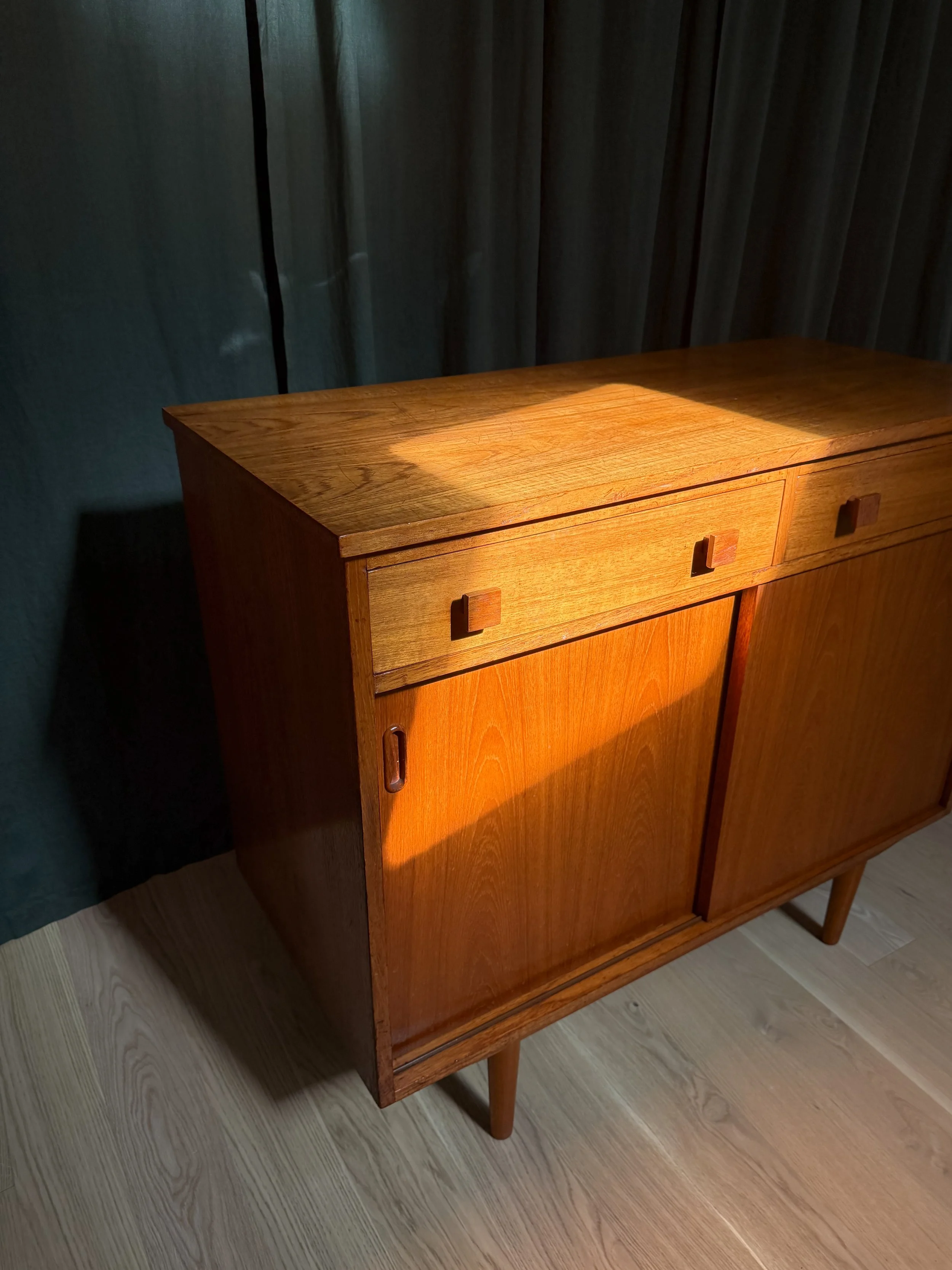 A wooden cabinet with sliding doors, illuminated by sunlight, standing on a wooden floor with a dark curtain in the background.