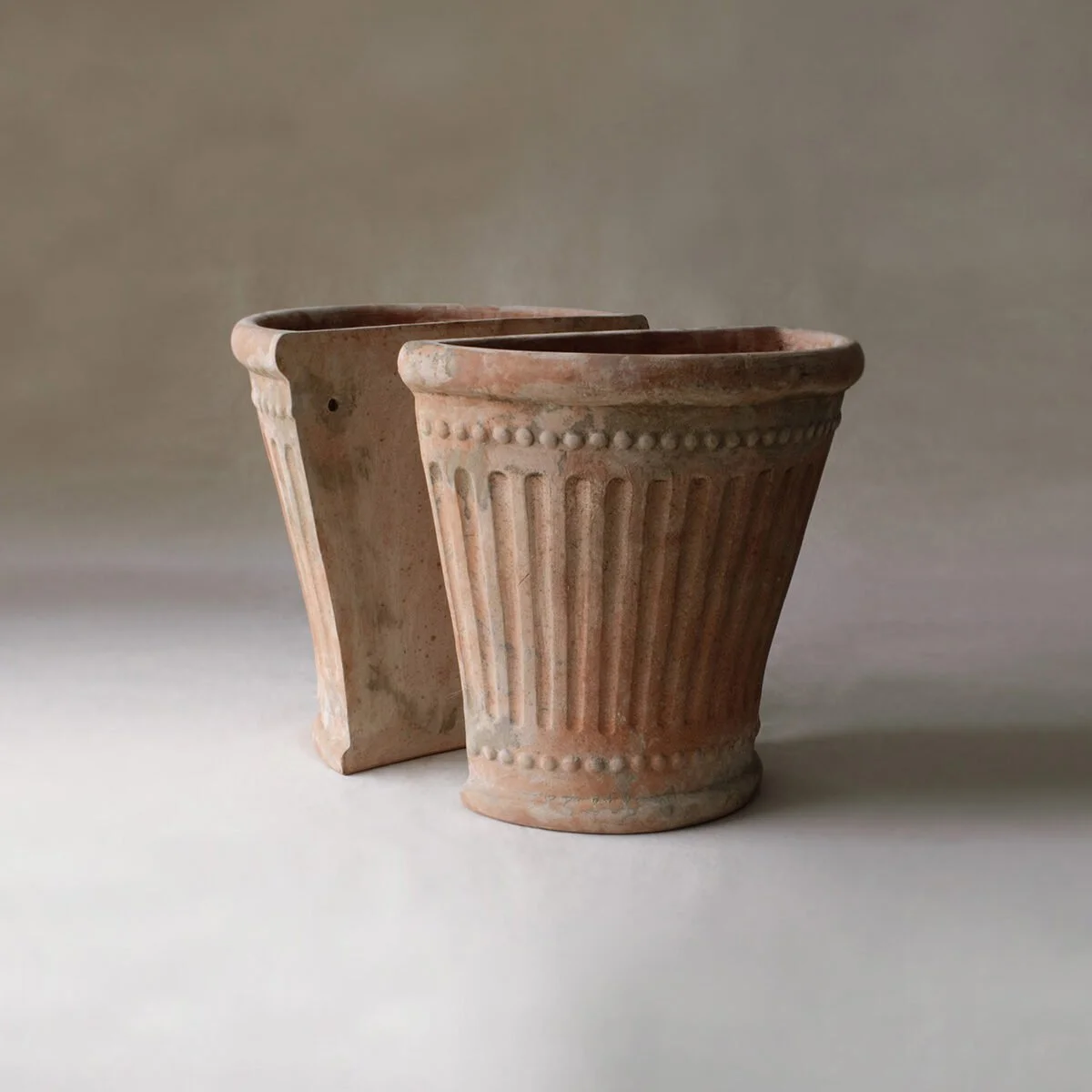 Two ancient terracotta flower pots with vertical fluted designs and decorative beaded patterns at the top, placed on a neutral surface against a plain background.