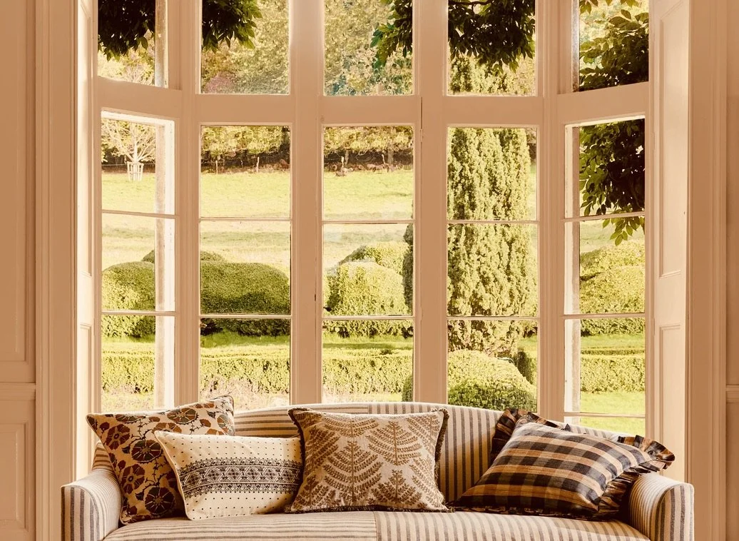 A bright living room with large bay windows overlooking a lush garden with various green shrubs and trees. A striped sofa with multiple decorative pillows is in front of the windows.