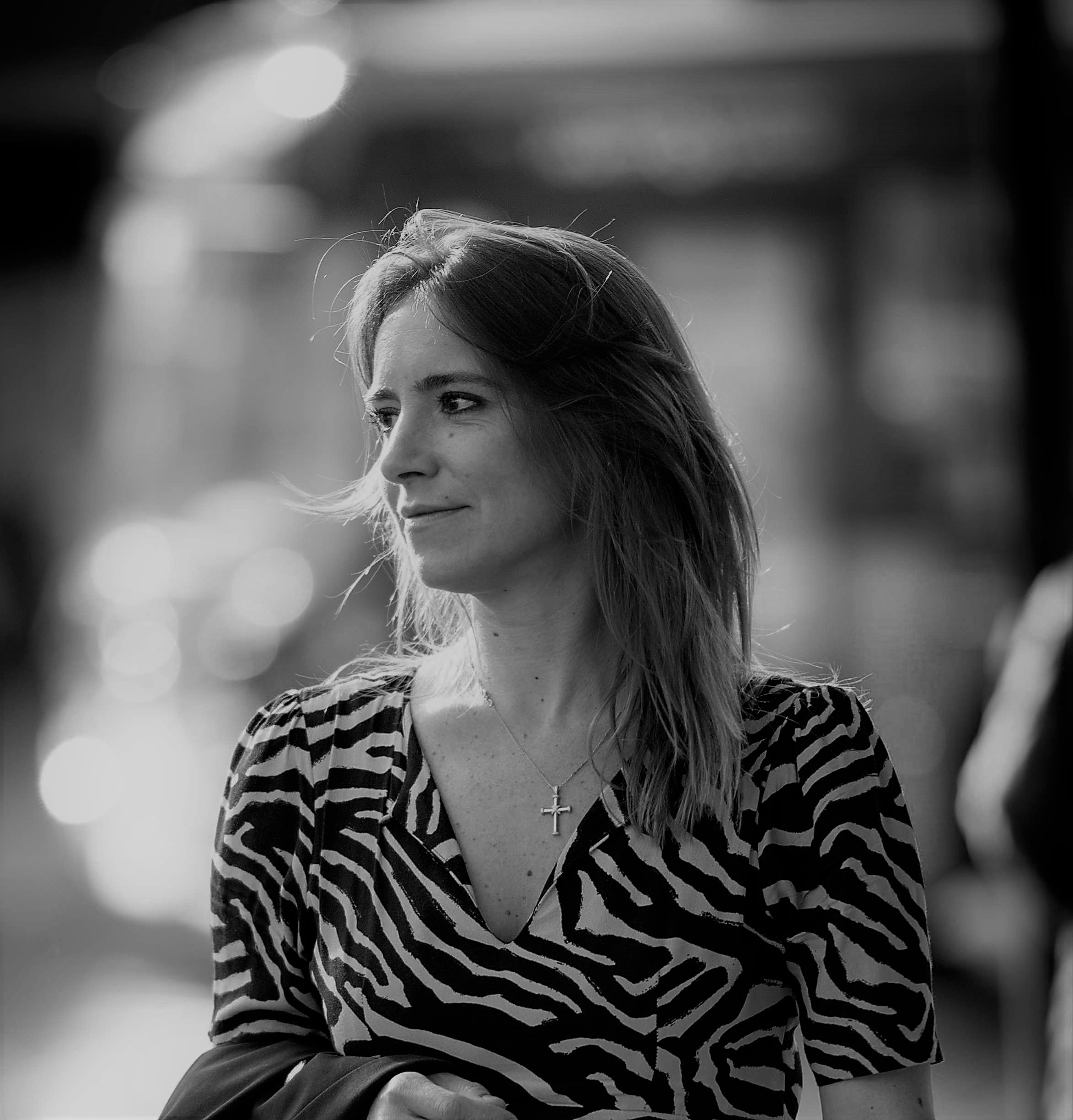 Black and white photo of a woman with long hair wearing a striped shirt, with a necklace that has a cross pendant. She is looking to the left with a neutral expression, and the background is blurred with light bokeh effects.