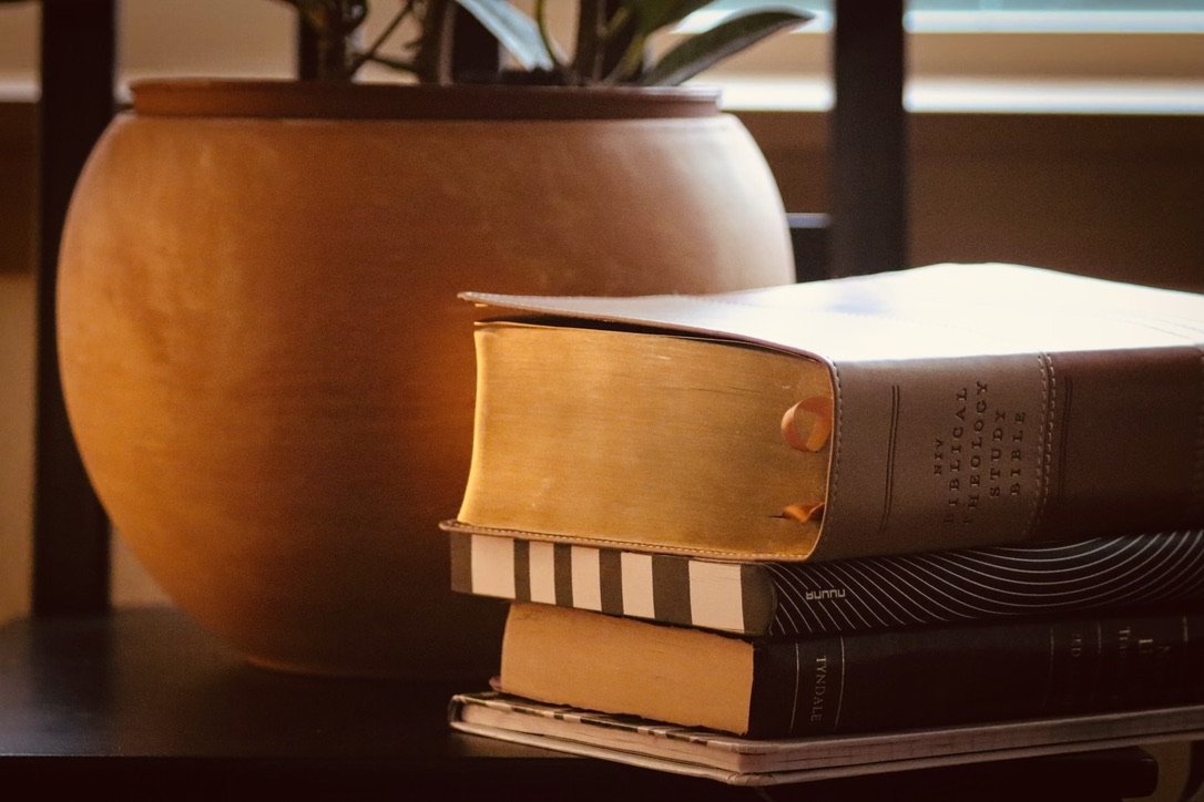 A stack of three books placed on a dark surface next to a large brown claypot. The top book shows the pages spread open, revealing a yellowish tone, with the title partially visible. The middle book has a black and white striped cover, and the bottom book is dark with visible text.