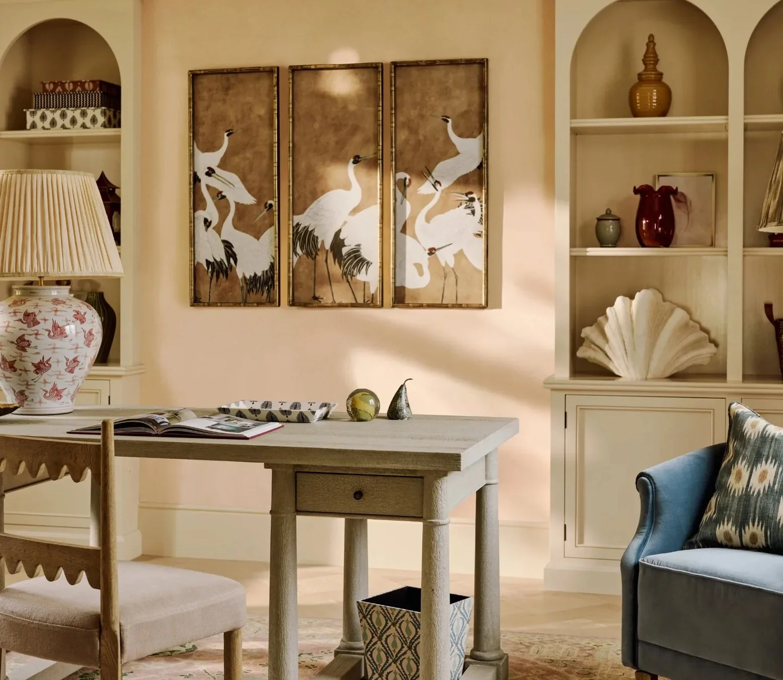 A cozy living room with a large beige wall featuring a triptych of white cranes on brown backgrounds. A wooden table with decorative objects, an open magazine, and a large ceramic lamp with a bird design on its base are in the foreground. White shelves with various vases, a seashell sculpture, and framed artwork are on the right side. A blue armchair with a pillow is partially visible on the right.