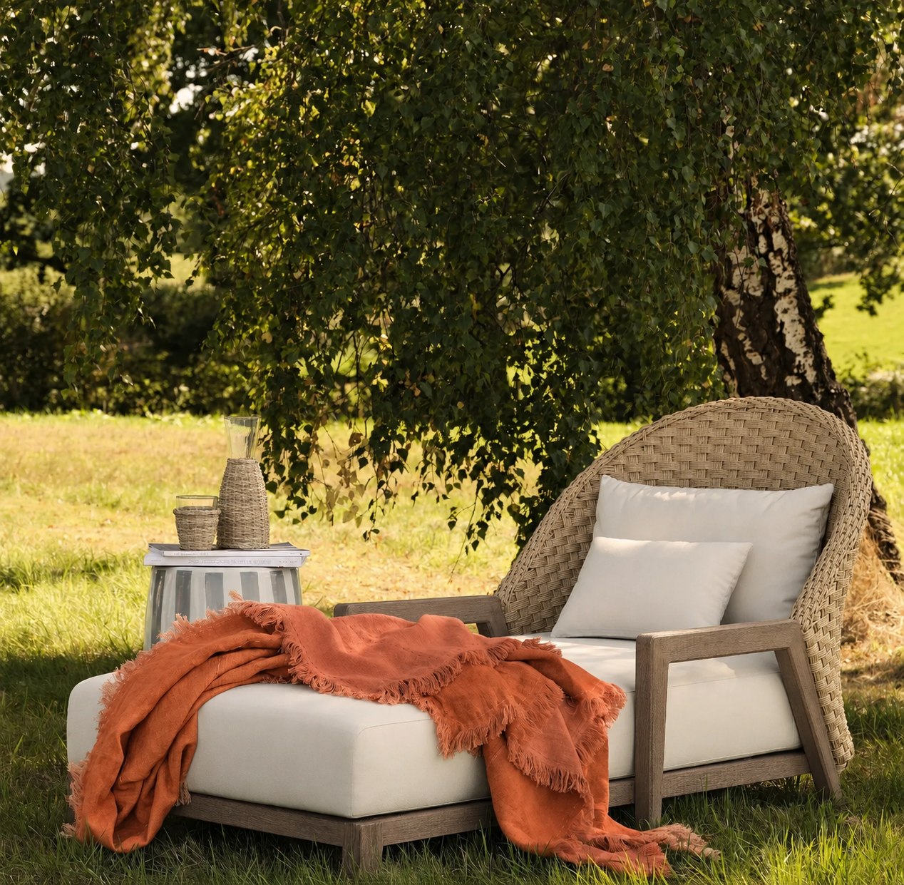 Outdoor lounge chair with white cushions and an orange throw blanket, positioned near a tree with lush green foliage, with a small side table holding a pitcher, glass, and woven containers.