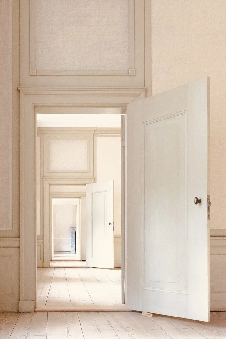 Multiple white doors open at different depths creating an infinite corridor effect in a bright room with light wood flooring and cream-colored walls.
