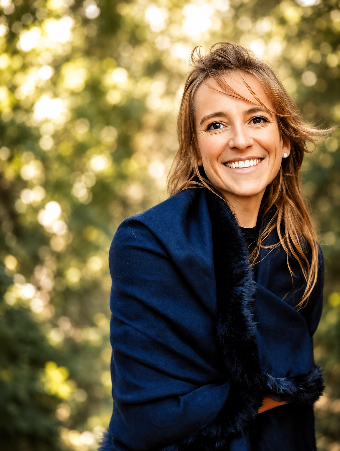 A woman with light brown wavy hair smiling outdoors, wearing a navy coat with a black fur collar, in front of a background of green trees and bright sunlight.