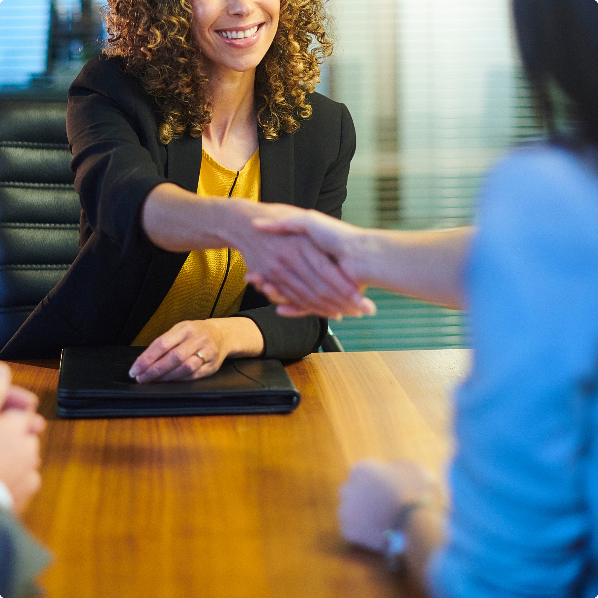 A woman with curly hair in a business suit greeting another person with a handshake in a conference room.