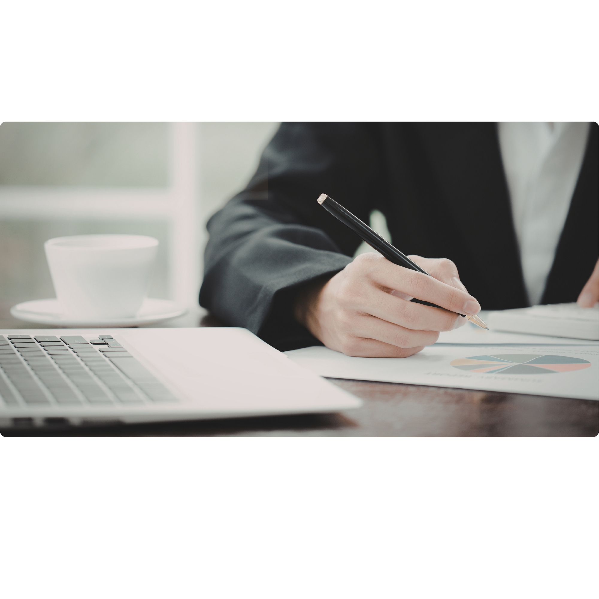 Person in business attire sitting at a desk with a laptop, holding a pen and working on documents, with a cup of coffee nearby.