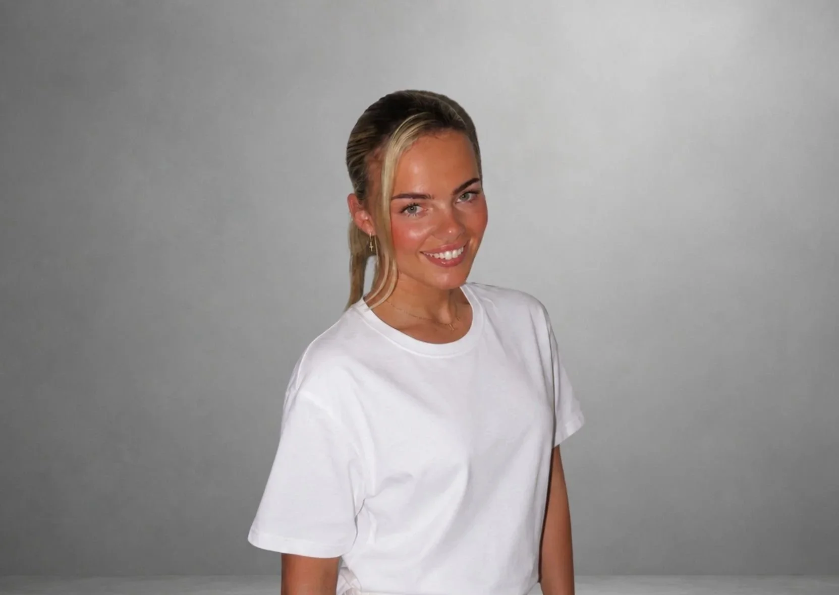 A woman with blonde hair styled in a loose wave, wearing a white t-shirt, smiling, with a light gray background.