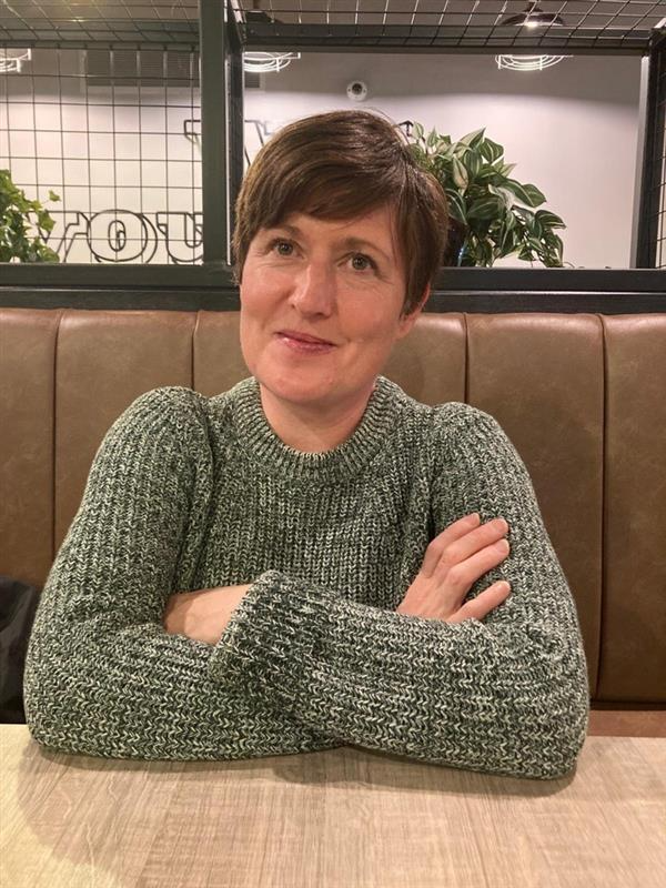 A woman with short brown hair, wearing a gray knit sweater, sitting at a table in a cafe or restaurant. She has arms crossed on the table and is smiling slightly, with indoor plants and a window behind her.