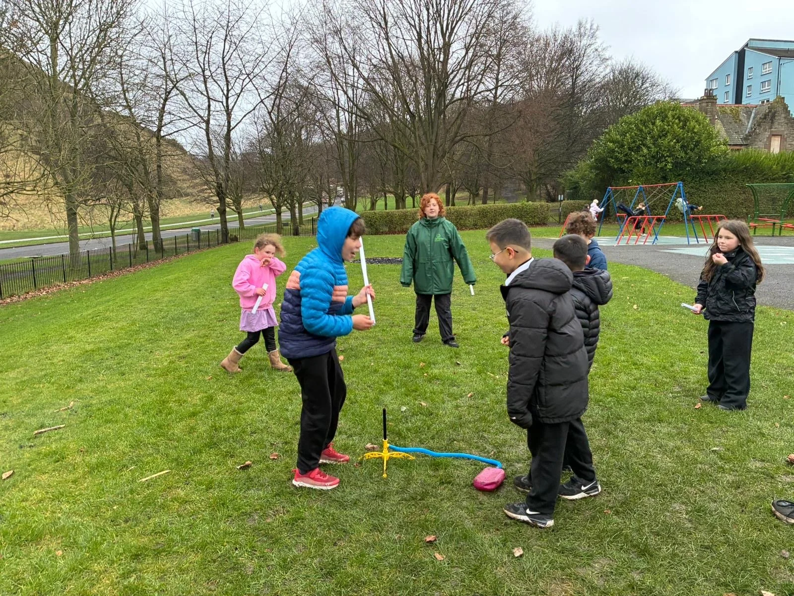 Children playing hopscotch and using skipping ropes outdoors on a grassy field in a park with trees and playground equipment in the background.