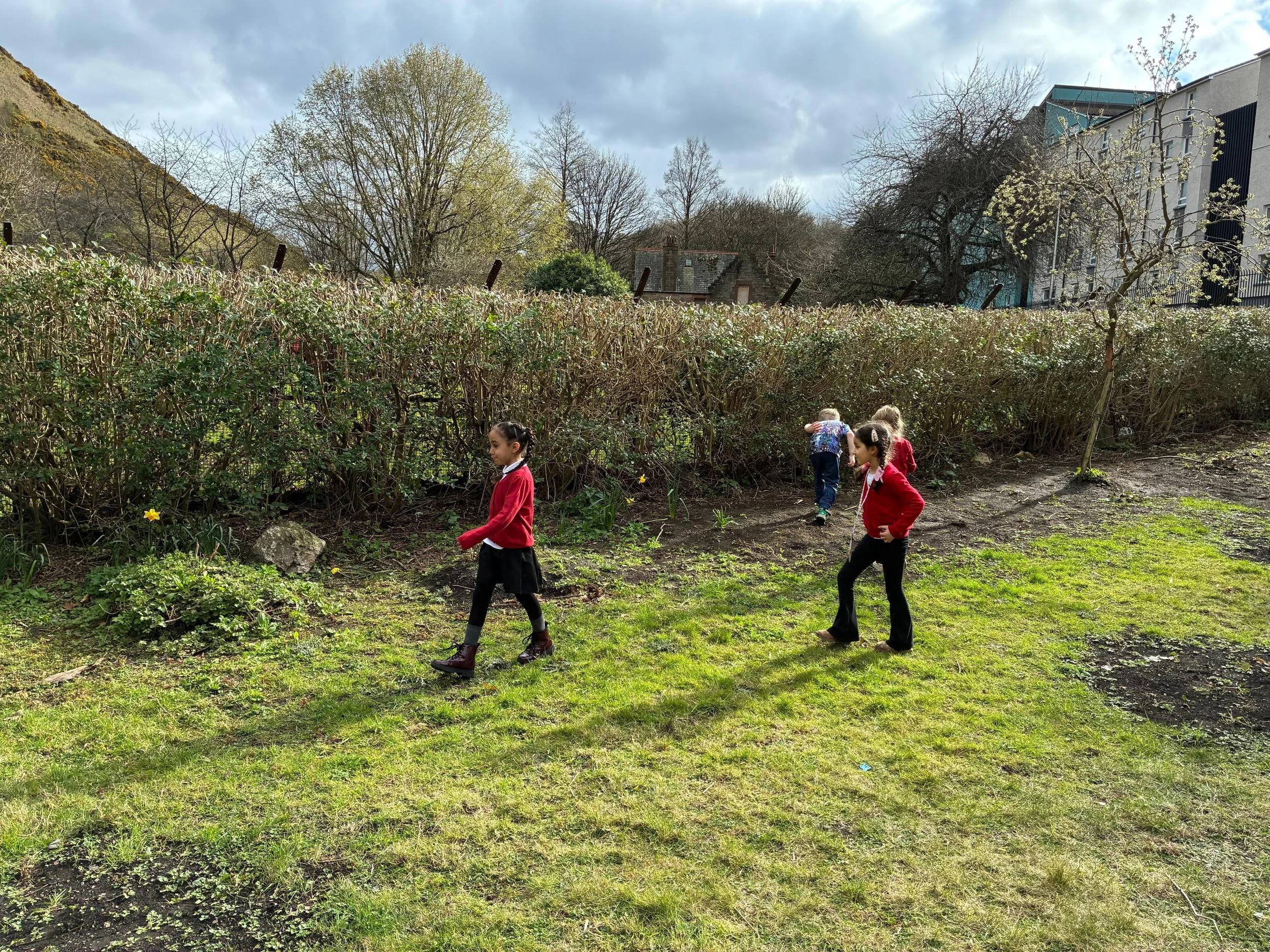 Children playing outdoors on grass near a hedge, with trees, houses, and a cloudy sky in the background.