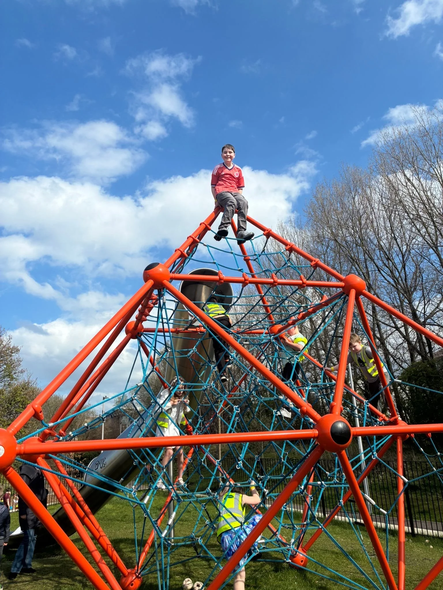 Children playing on an orange and blue climbing playground structure outdoors on a sunny day with a blue sky and scattered clouds, with trees in the background.