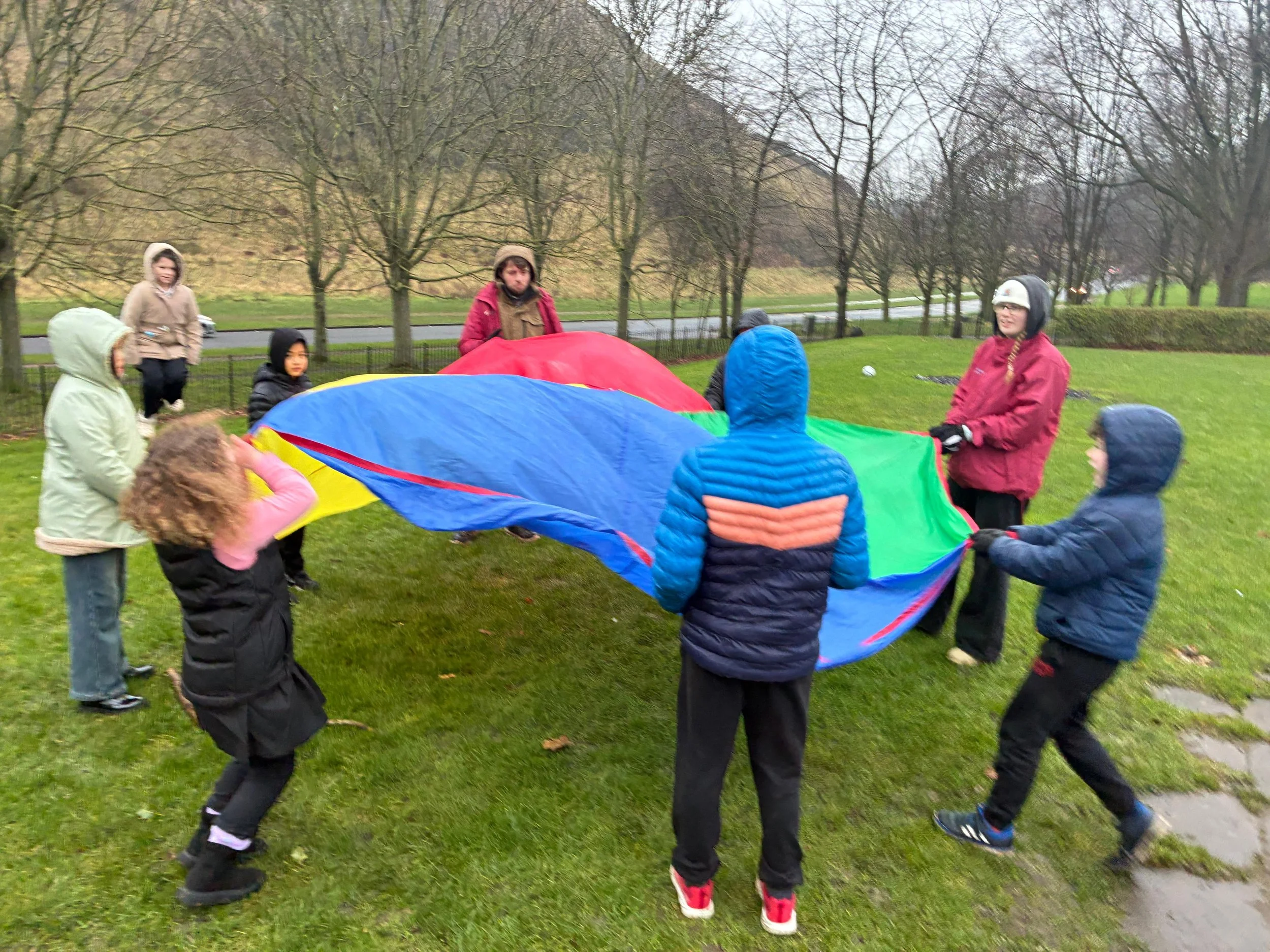 Group of children and adults outdoors playing with a colorful parachute on a grassy area with trees and a road in the background.