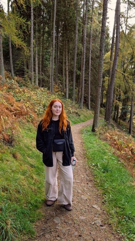 A young woman with long red hair standing on a dirt trail in a forest with tall trees and green foliage.