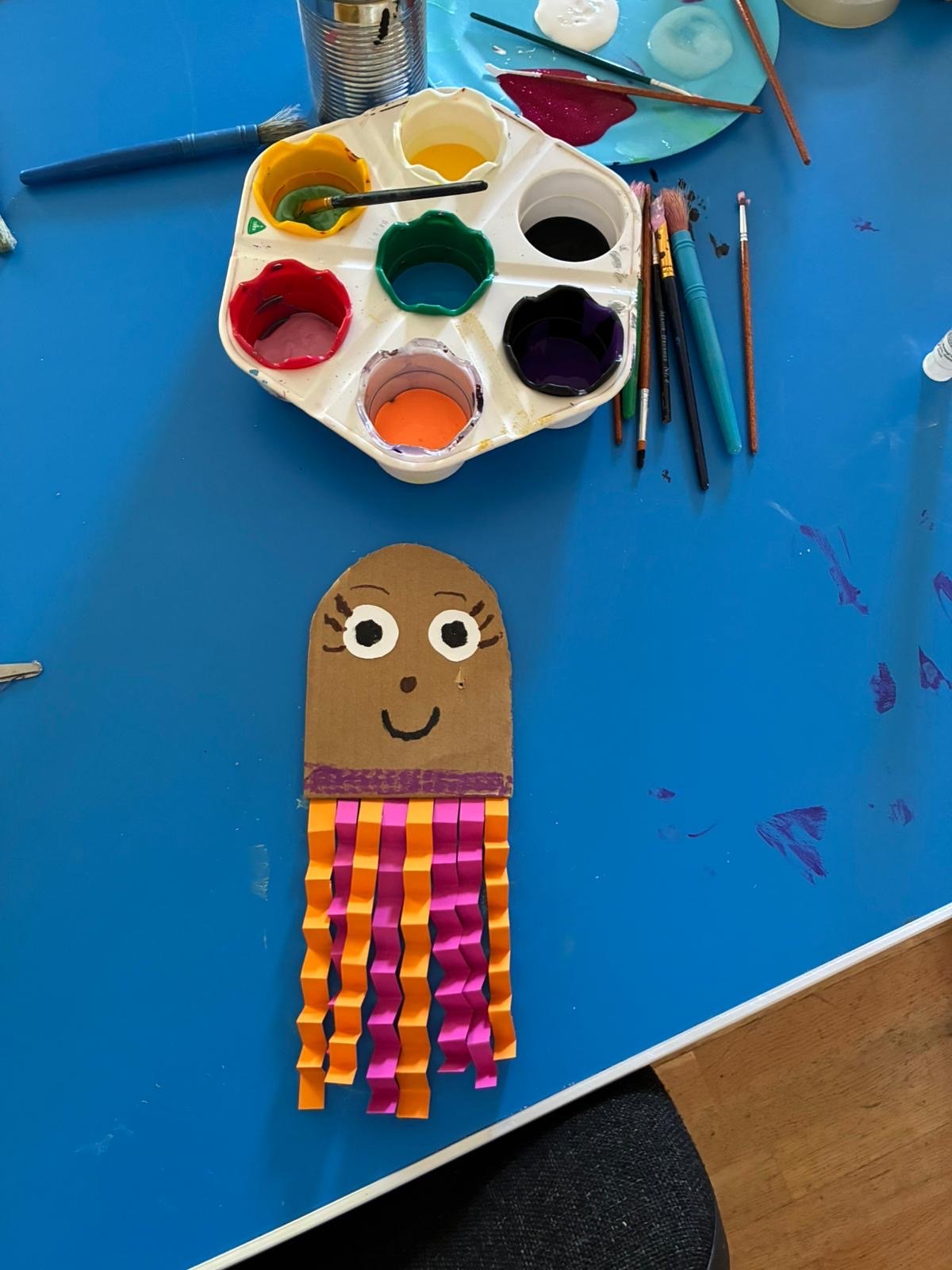 A craft project of a popsicle stick octopus with a cartoon face, drawn eyes and mouth, topped with a paper head, on a blue table with paint supplies.
