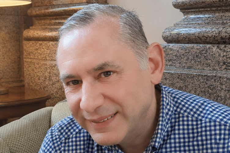 A smiling middle-aged man with short gray hair, wearing a blue checked shirt, sitting on a beige couch with decorative stone walls in the background.