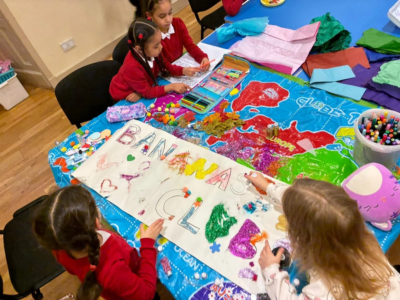 Children participating in a craft activity at a table covered with art supplies, decorations, and a large collaborative poster that says 'BANANA SPLIT' on it.