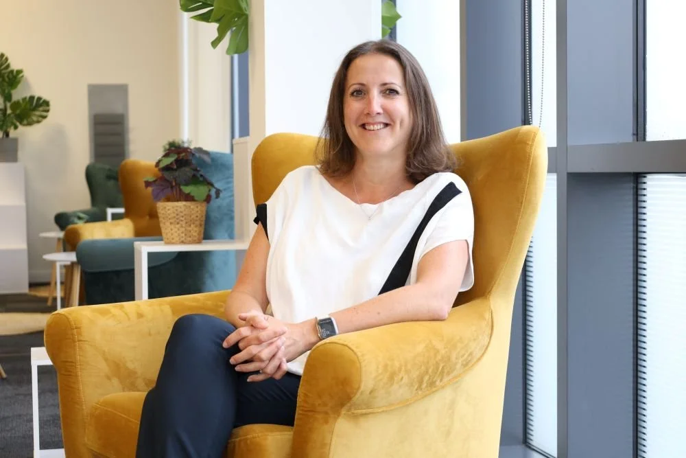 Woman sitting in a yellow armchair in a modern, brightly lit lounge area with potted plants and colorful chairs in the background.