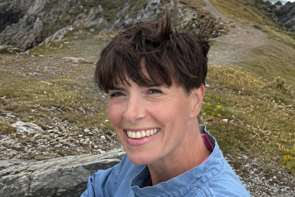 A woman smiling outdoors in a mountainous area with rocky terrain and grass.