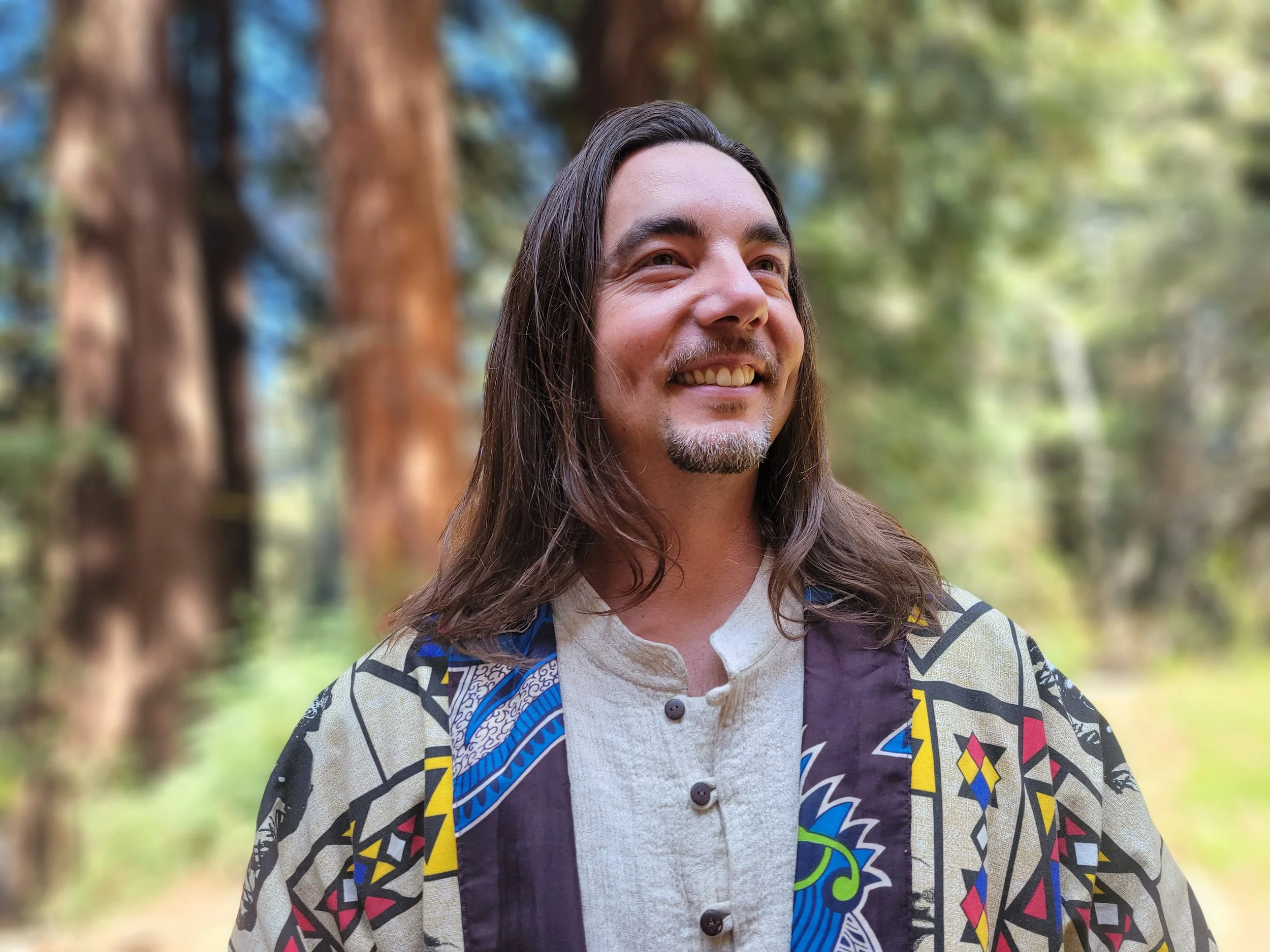 A man with long brown hair and a goatee, smiling, in an outdoor forest setting, wearing a beige button down collar-less shirt, and a colorful kimono jacket with red, yellow, and black african art patterns, and a black and blue accent lapel. 