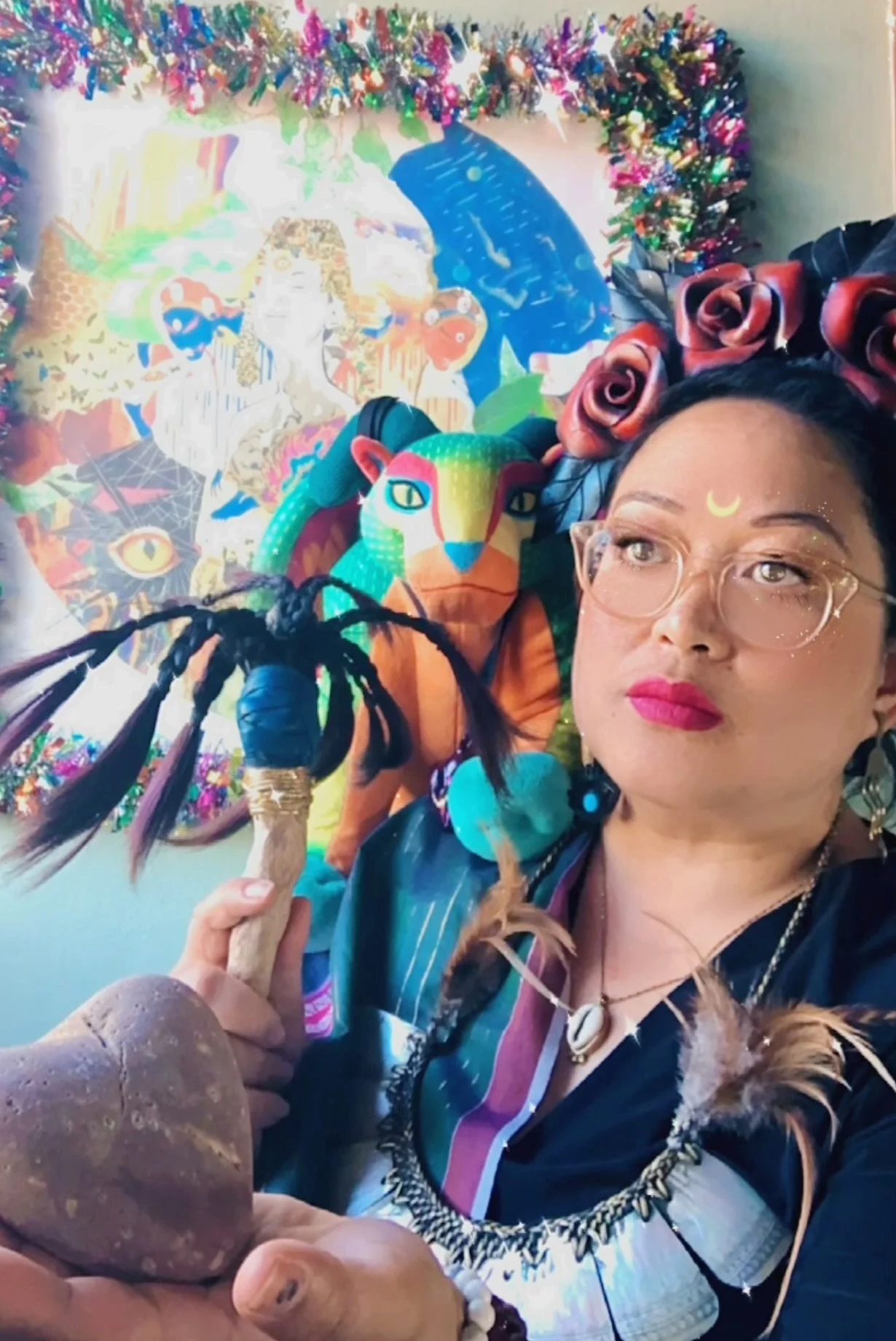 A Filipina woman with glasses, pink lipstick, and a crescent moon painted on her forehead holds a dreamcatcher in front of a colorful background with vibrant, artistic mural with mythical and animal themes, and she is wearing a pink flower crown.