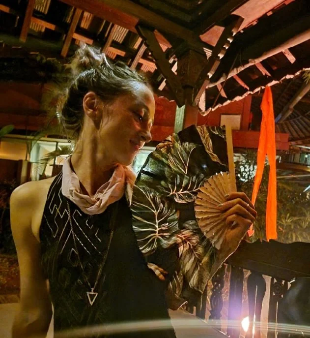 A woman in a black dress with gold geometric line designs, looking over her left shoulder, holding a decorative asian-style fan covering her left shoulder, in a dimly lit, rustic setting with wooden beams and vibrant orange and red accents.