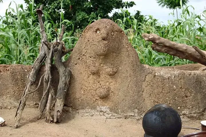 An earthen figurine with cowry shell eyes, nubs of earth marking a nose, breasts, and a bulge around the navel. A low wall extends out from both sides, and a group of sticks is stacked on the left side. It appears to be a type of shrine.