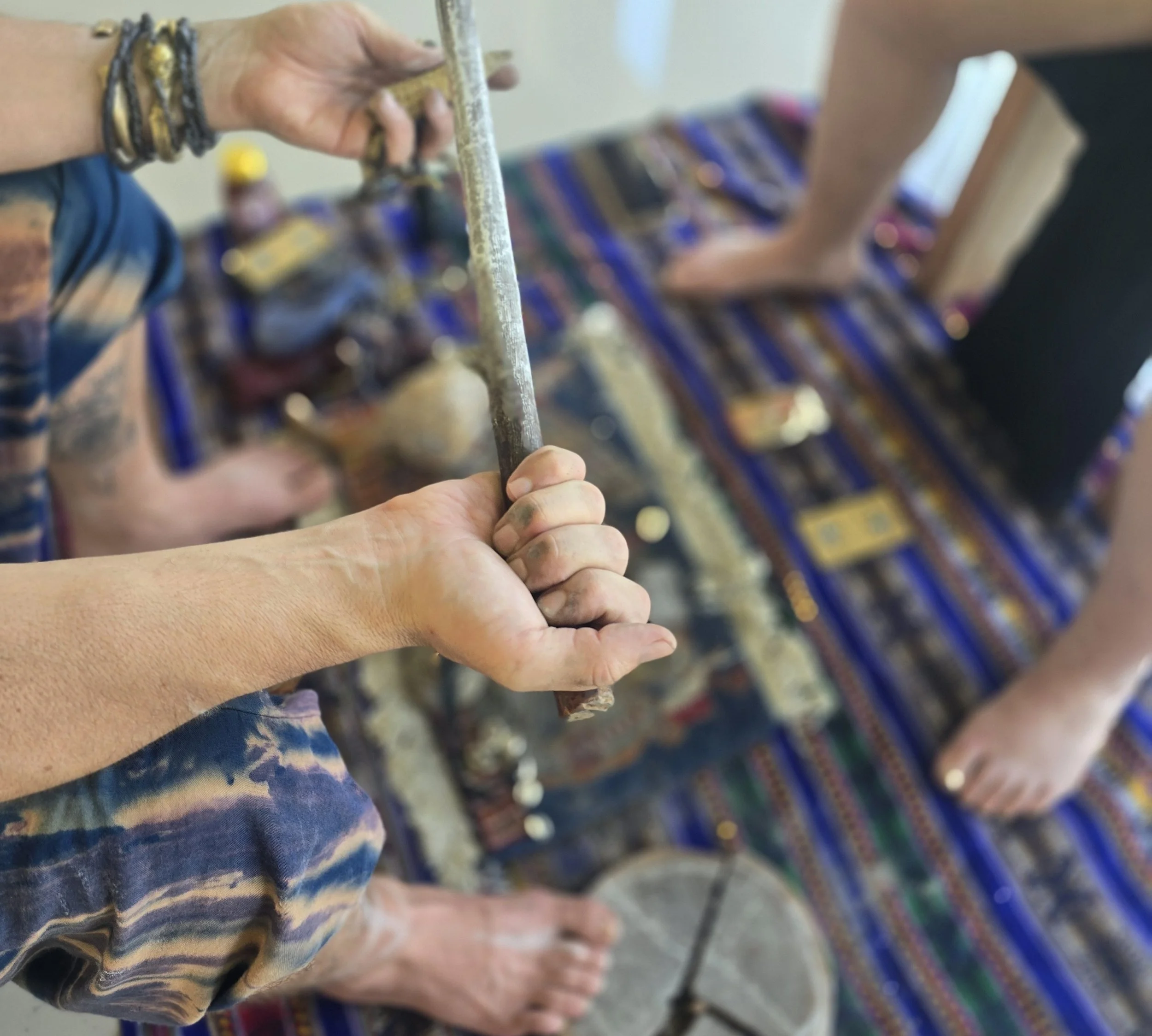 A man's hand smudged with ash, holds a stick with chalk markings. His opposite hand is holding a small corn cob, and his wrist is adorned with many metal bracelets. The background, blurred, shows a decorative blue cloth covered with sacred items.
