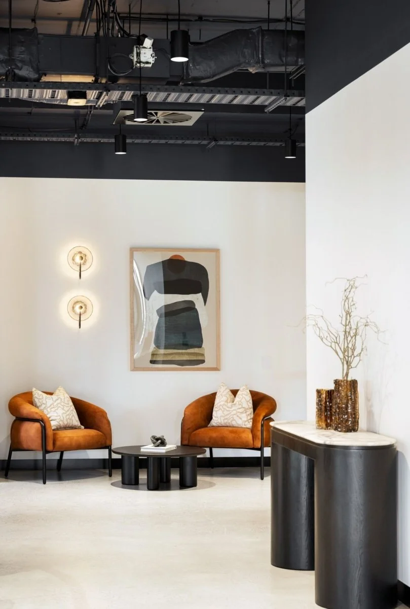 A modern lobby with two orange velvet armchairs, a black round coffee table, abstract artwork on the wall, and decorative lighting on a white wall. There is a side table with dried branches in vases.