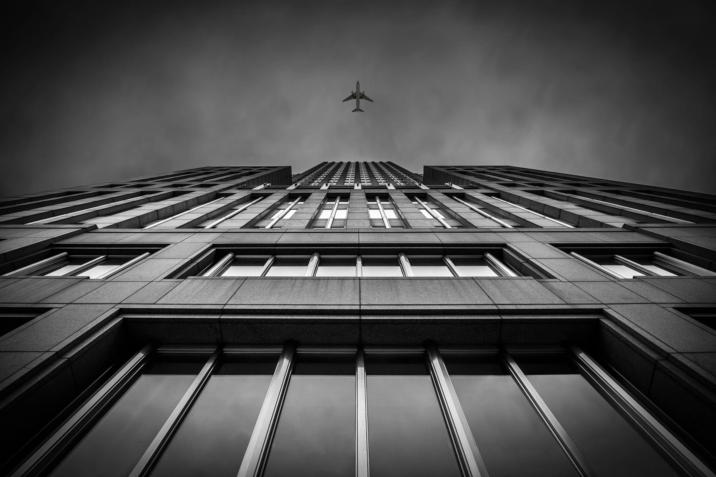 Low-angle view of a tall modern building with windows and metal framing, with an airplane flying overhead against a cloudy sky.