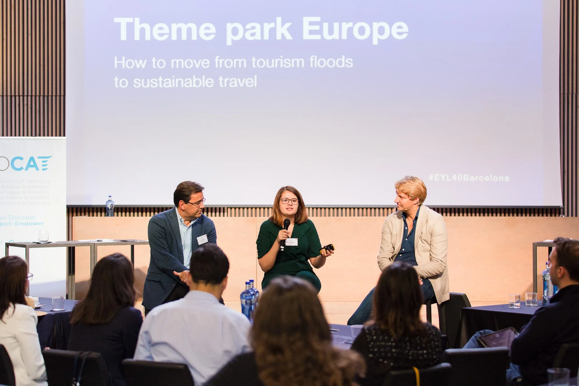 European Young Leaders panel discussion titled 'Theme park Europe' with Samantha Keon speaking into a microphone and two men listening, in front of a presentation slide on sustainable travel and tourism.
