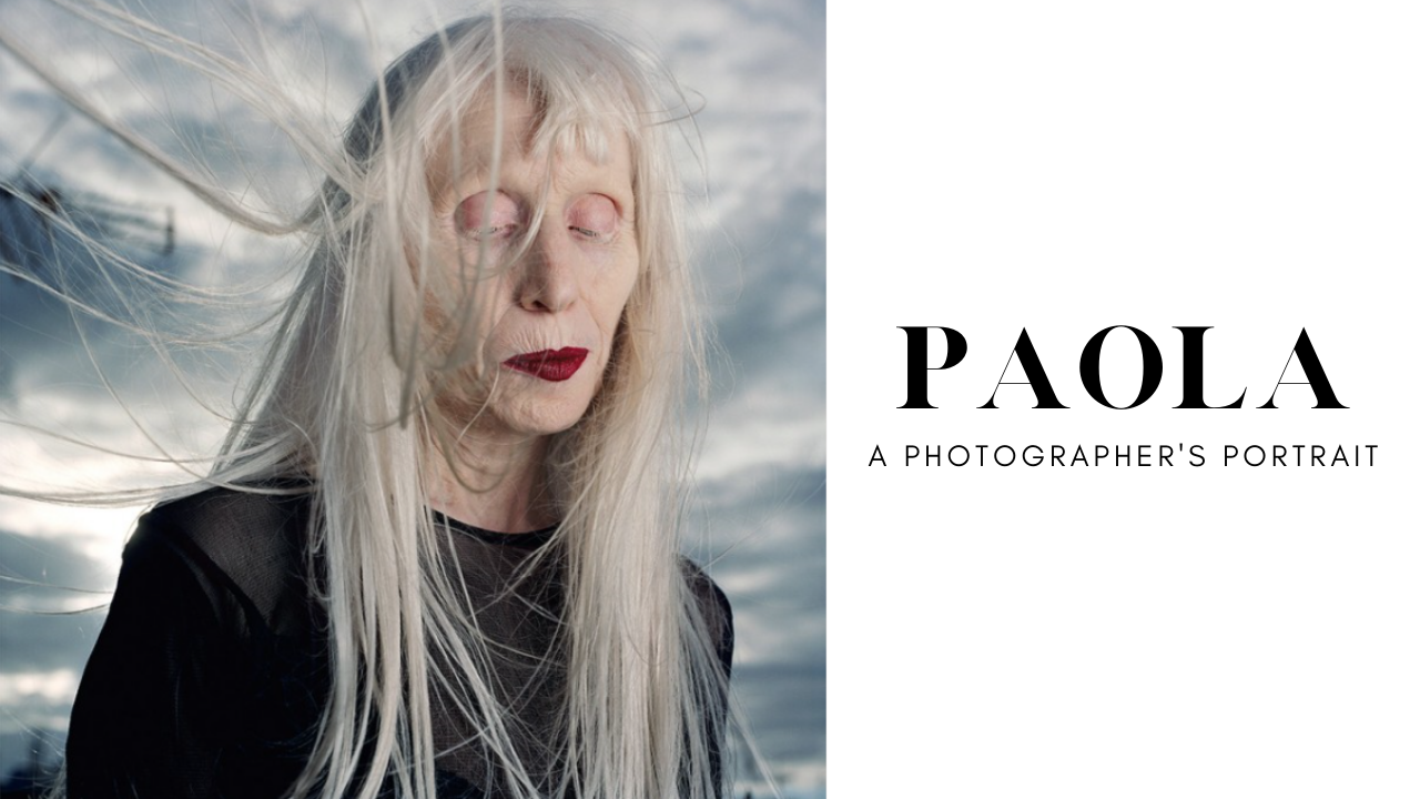 A portrait of an elderly woman with long blonde hair outdoors, wind blowing her hair across her face, against a background of cloudy sky, wearing a black top. Poster for the documentary short Paola