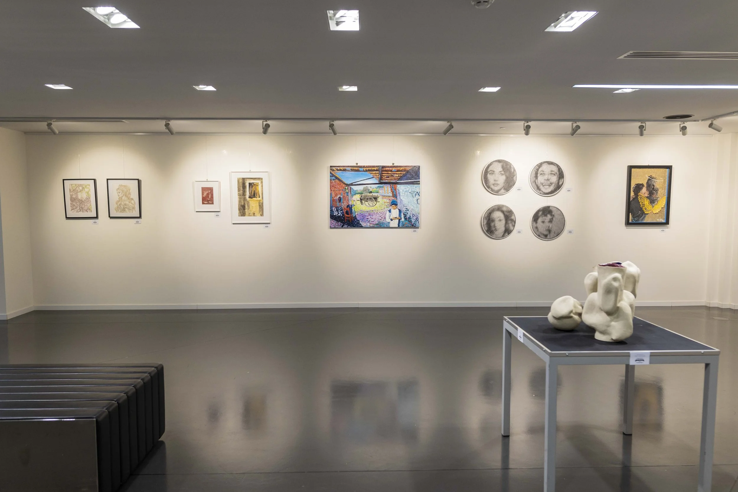 Une galerie d'art avec plusieurs œuvres accrochées au mur, dont des dessins, peintures, et photos. Un socle en métal avec une sculpture abstraite blanche au centre de l'espace.