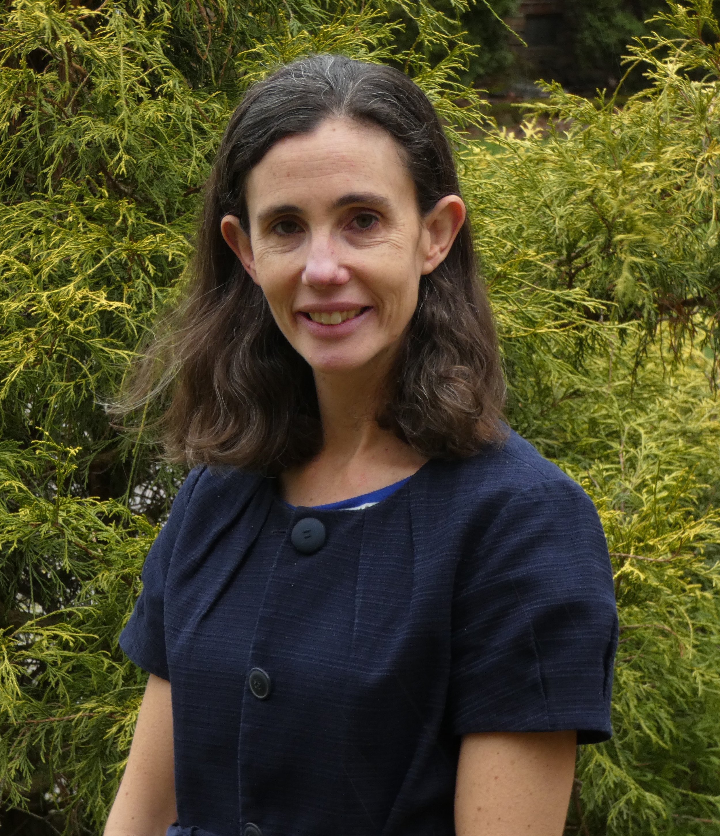 A woman with shoulder-length brown hair in a blue short-sleeved shirt standing outdoors in front of green foliage.