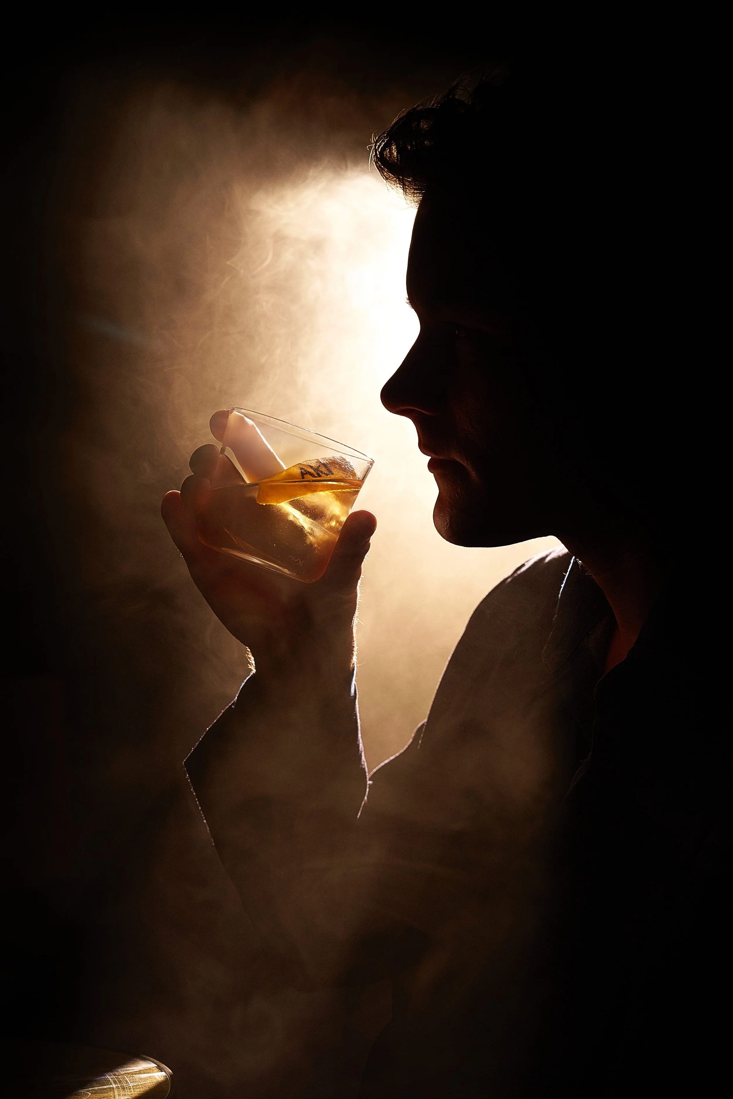 Silhouette of a man holding a glass of whiskey with ice, drinking in a dimly lit environment with a warm backlight.