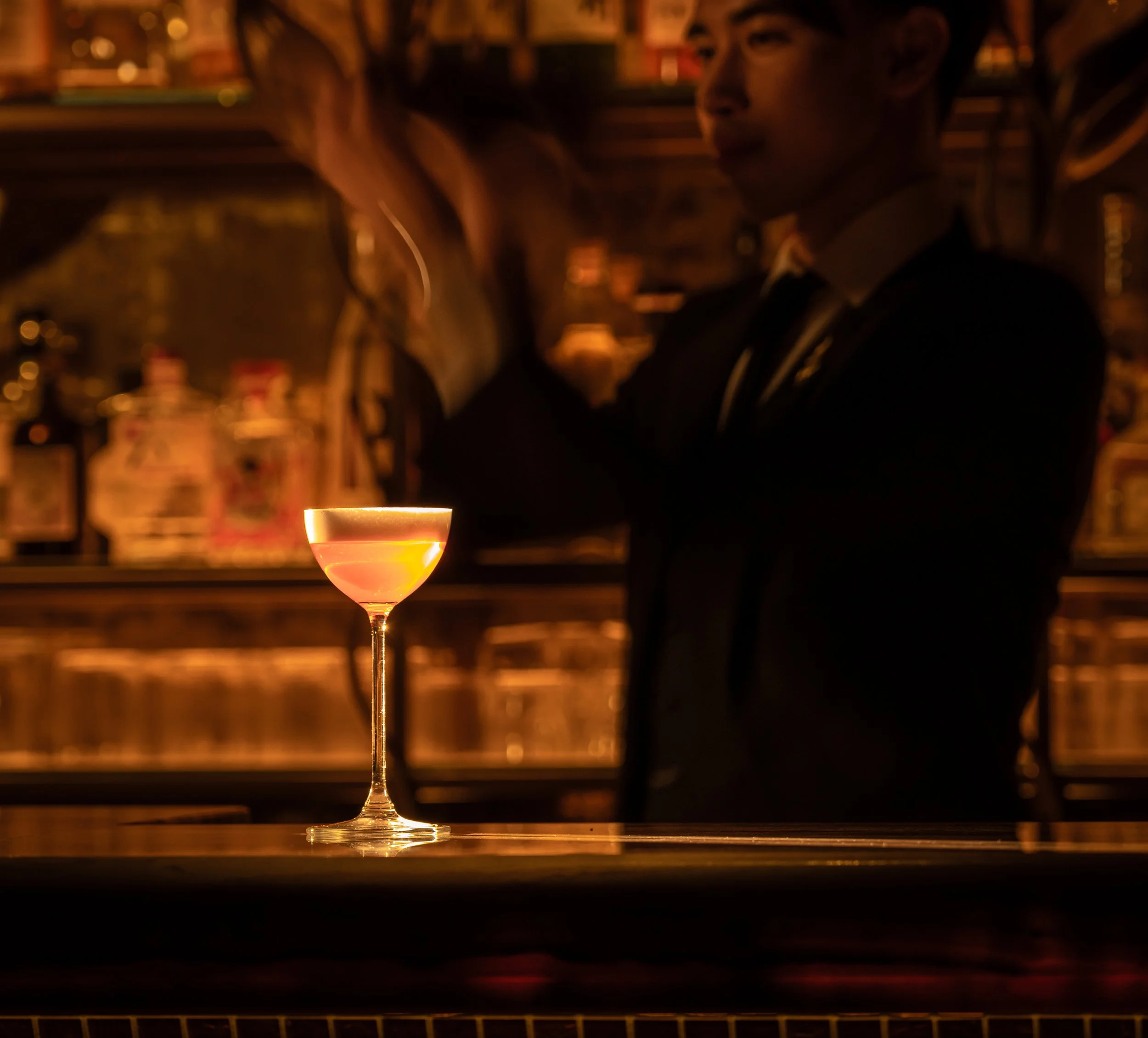 A cocktail glass filled with a pink-colored drink sitting on a bar counter with a bartender and a woman in the background.