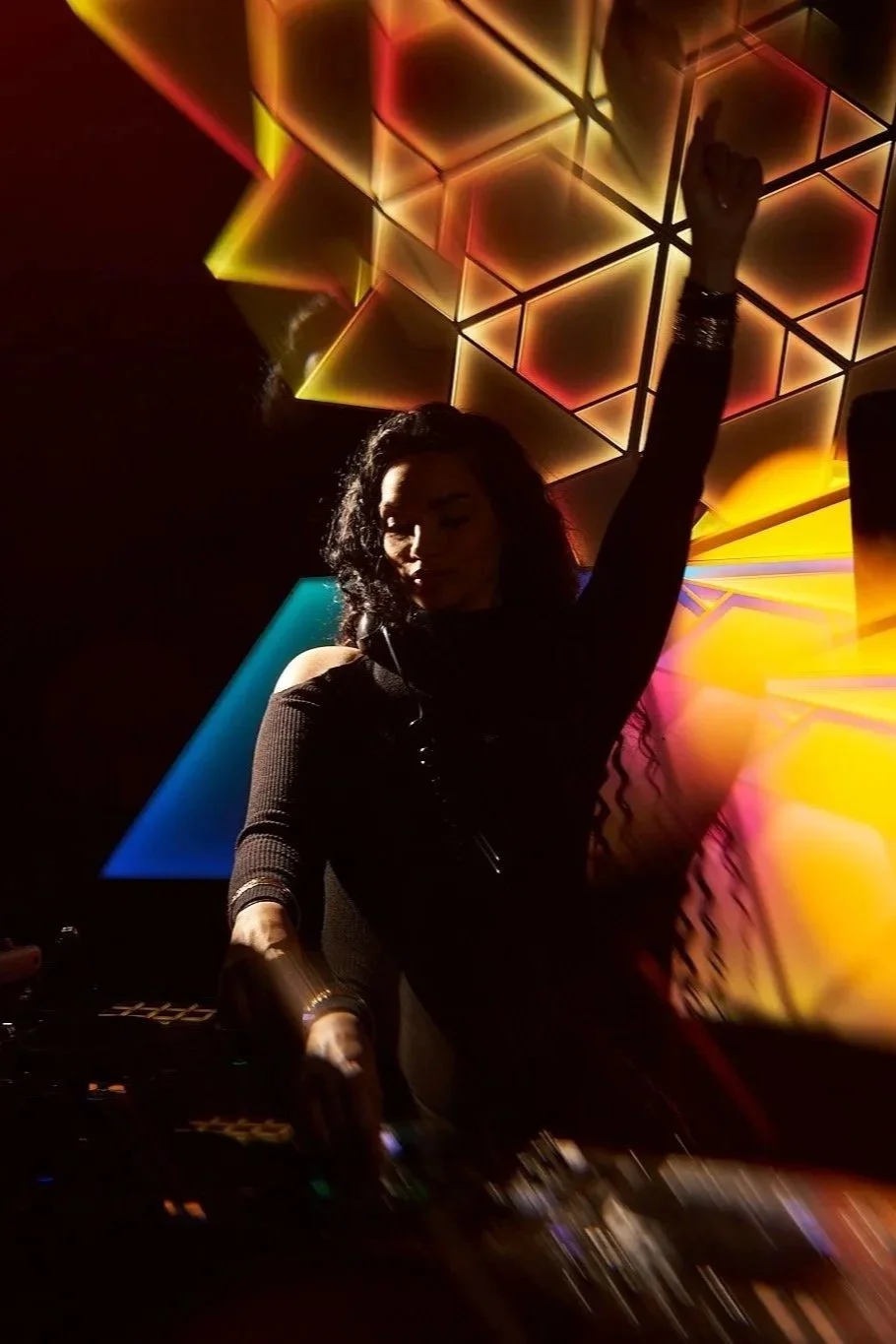 A woman with curly dark hair DJing in a nightclub with colorful LED light panels in the background, raising one arm.