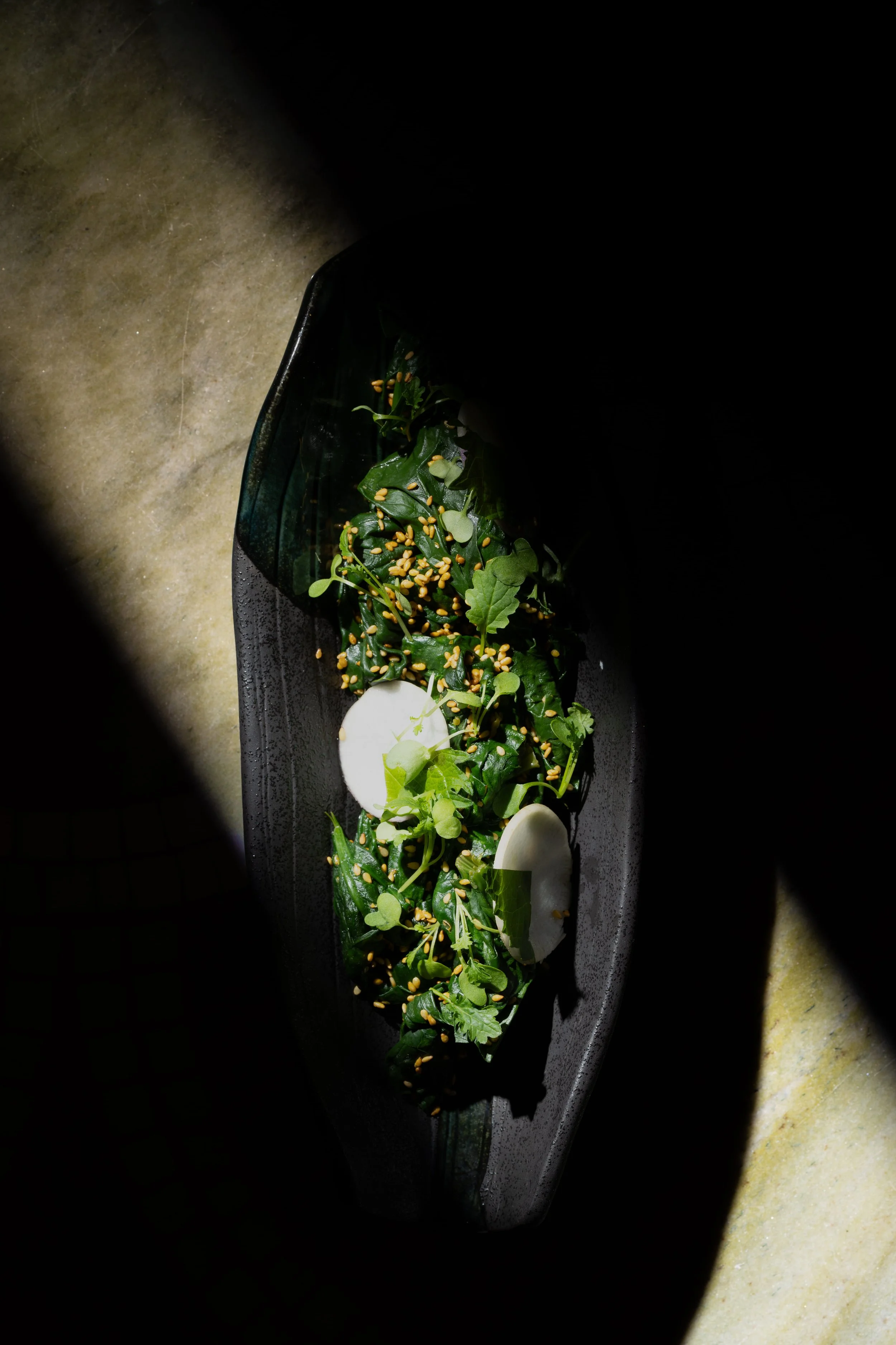 Salad with leafy greens, slices of radish, and sesame seeds on a black plate with dark lighting.