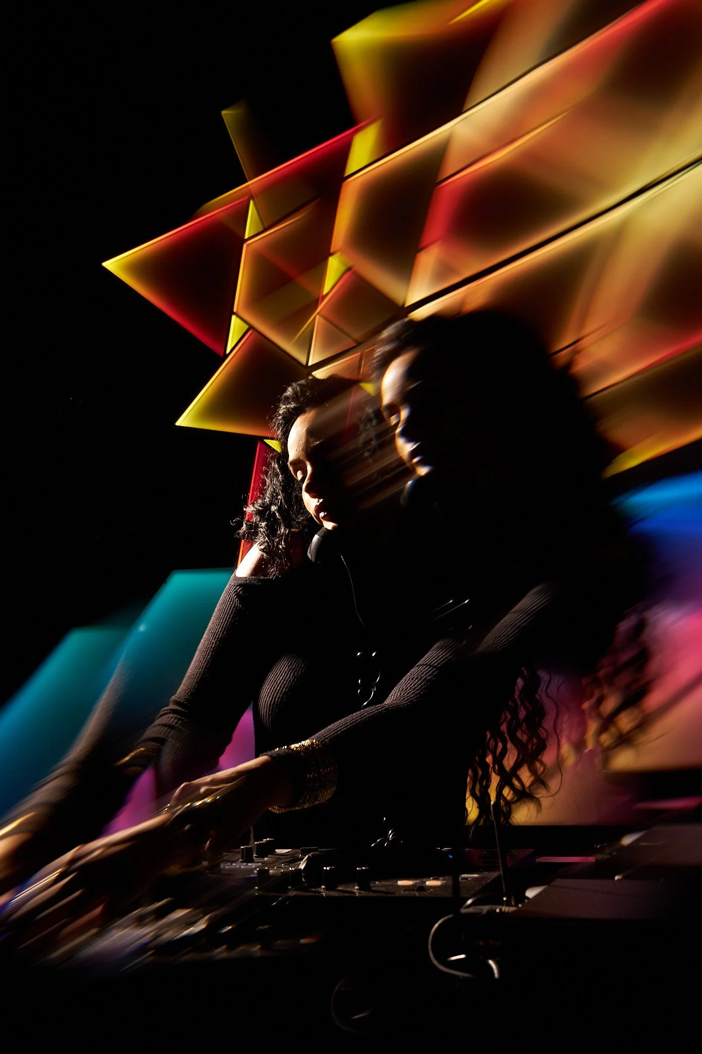 A female DJ is performing at a nightclub with colorful, geometric lights creating a streaking motion effect.